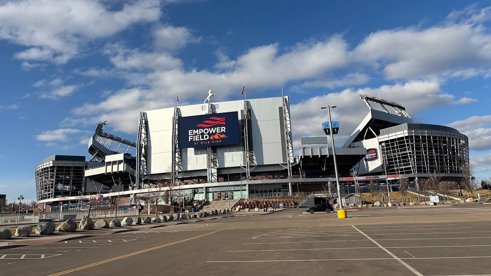 Fachada del estadio Empower Field at Mile High con su pantalla gigante y un caballo blanco sobre el techo, bajo un cielo azul con nubes.