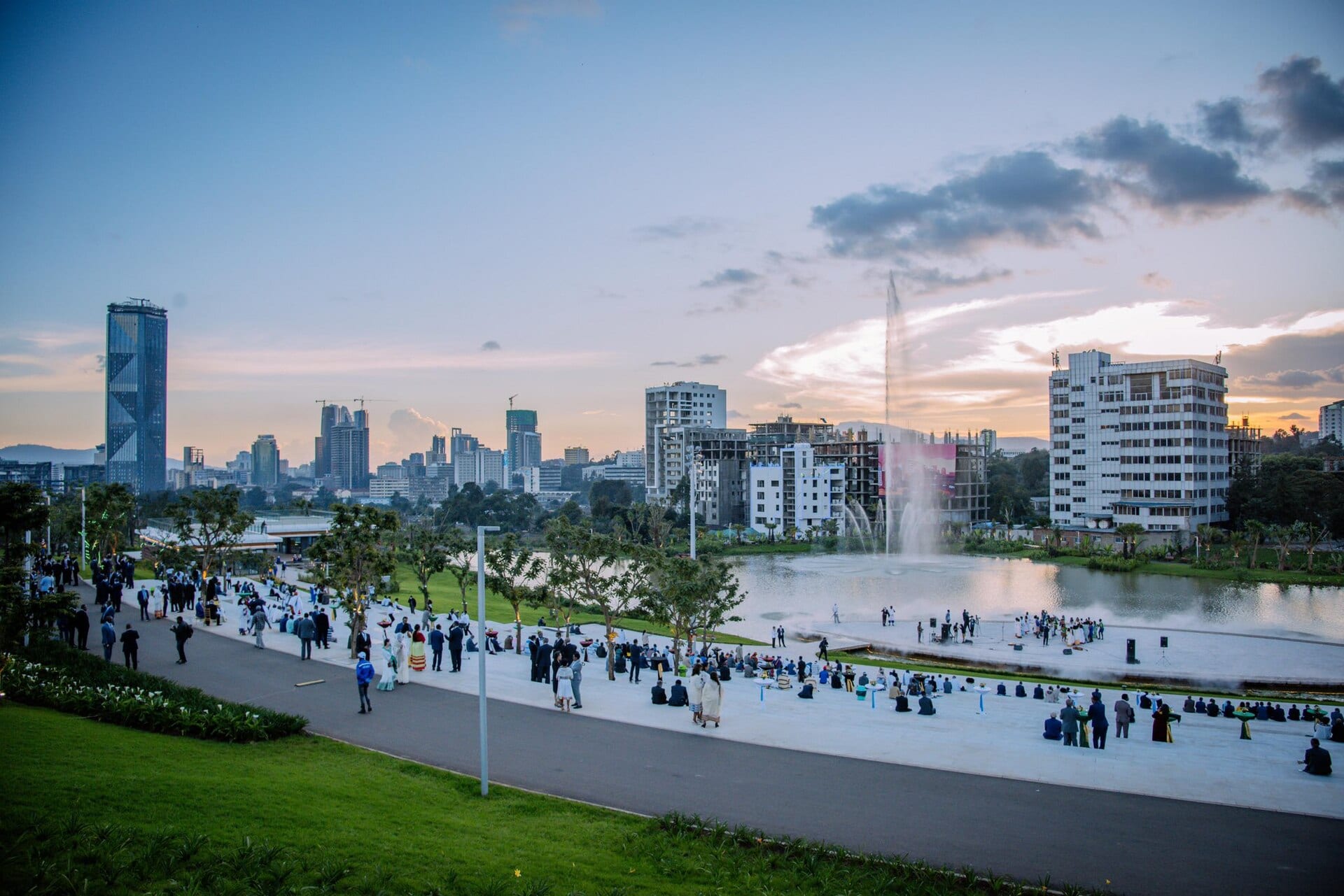 A lakeside park with a fountain, people gathered on the esplanade and tall buildings in the background under a sunset sky.