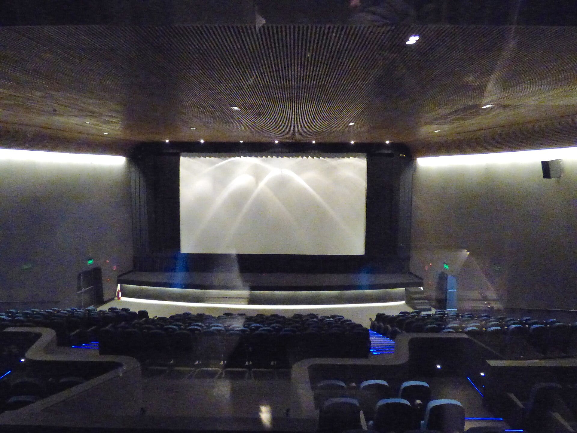 Sala de cine vacía vista desde la cabina de proyección, con la pantalla iluminada al fondo y filas de butacas oscuras.
