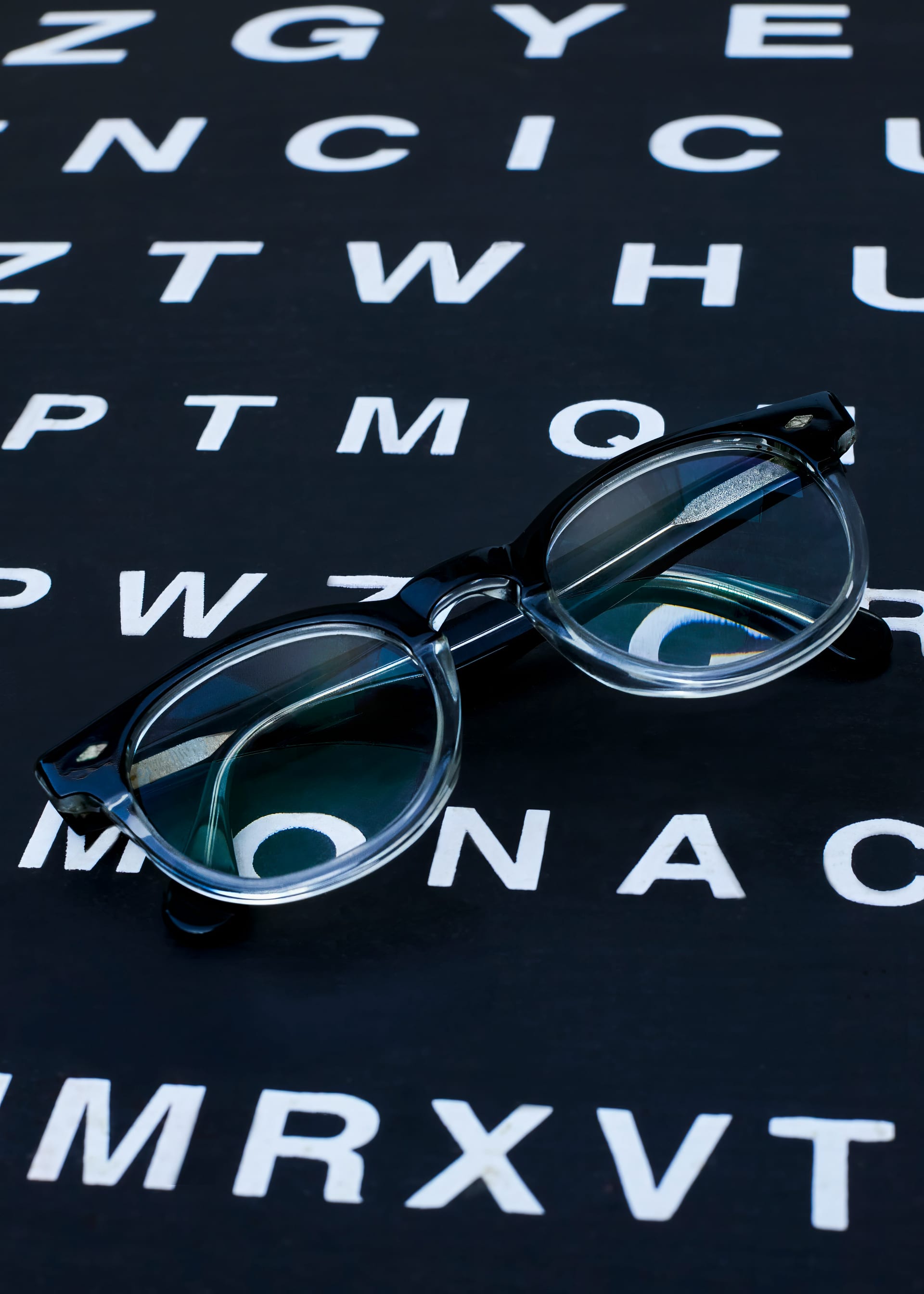 Unos anteojos de armazón negro con bordes transparentes descansan sobre una tabla de letras blancas en fondo oscuro, como las de un examen de la vista.
