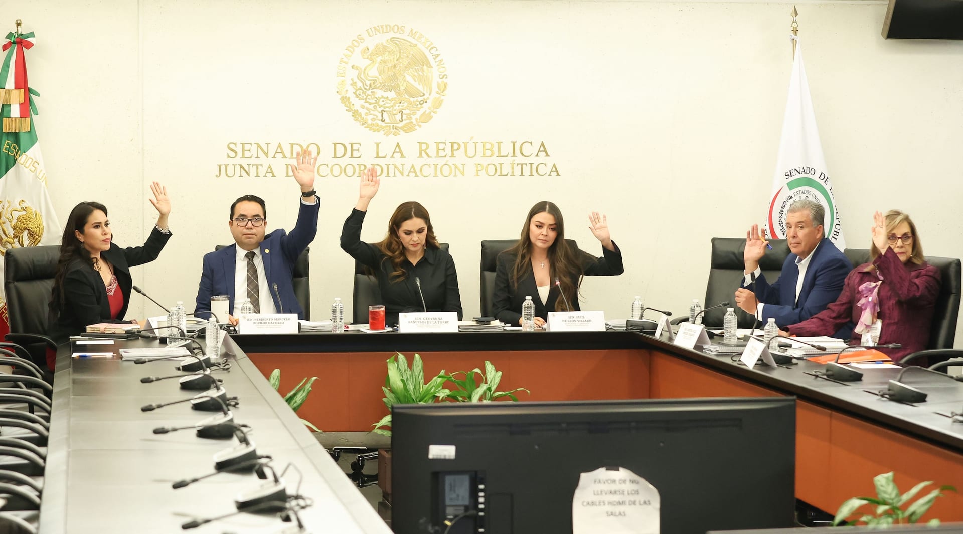Seis senadores y senadoras levantan la mano para votar en una sala de comisiones, bajo el letrero de la Junta de Coordinación Política del Senado.