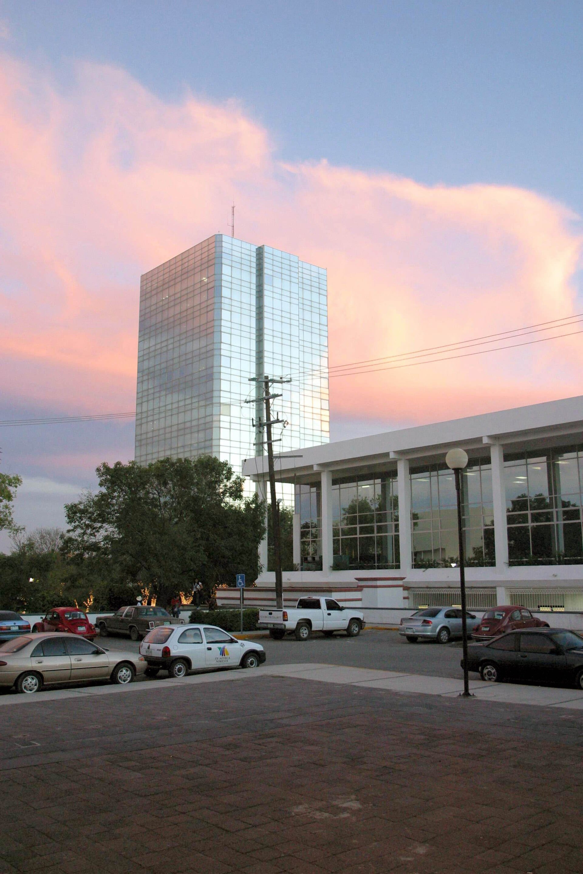 Una torre de cristal y un edificio blanco de oficinas públicas al atardecer, con autos estacionados al frente.