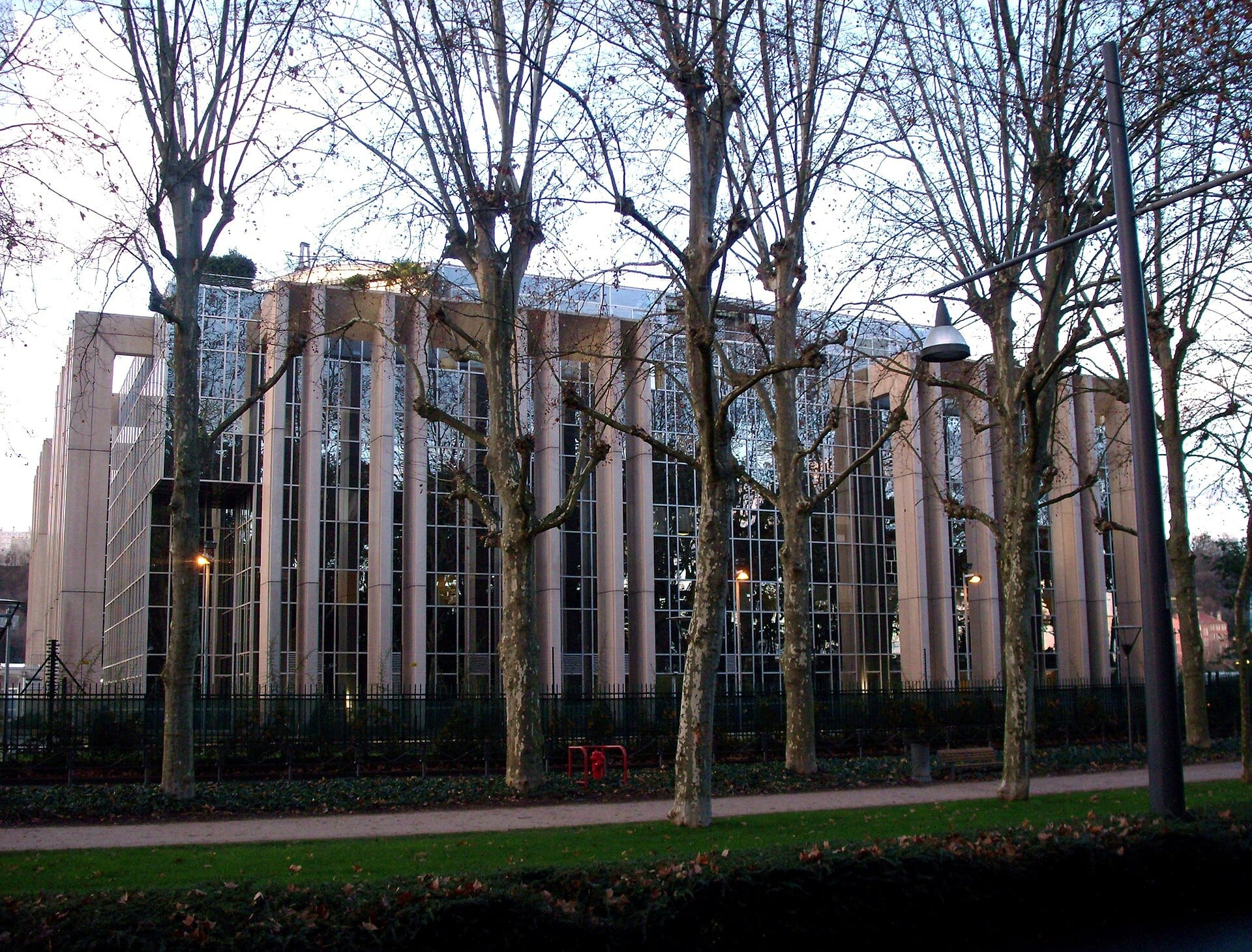 Edificio de cristal con columnas claras, visto entre árboles sin hojas, con un camino y césped en primer plano.