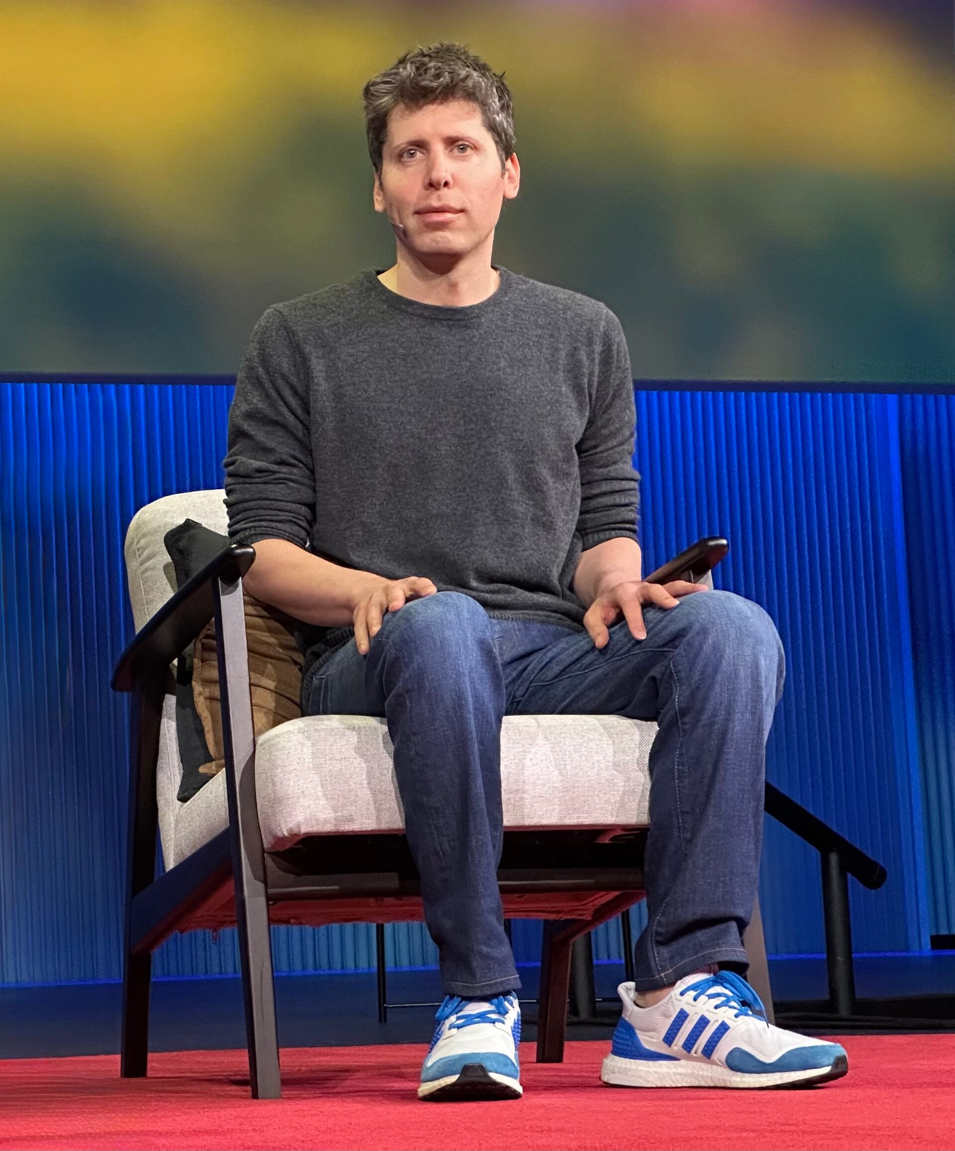 Sam Altman habla de pie con un micrófono de mano frente a un fondo oscuro en un escenario.