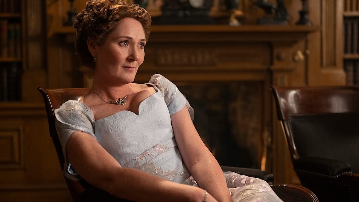 A woman in a light blue dress and necklace sits on a wooden chair in a paneled library with bookshelves and a fireplace behind her.