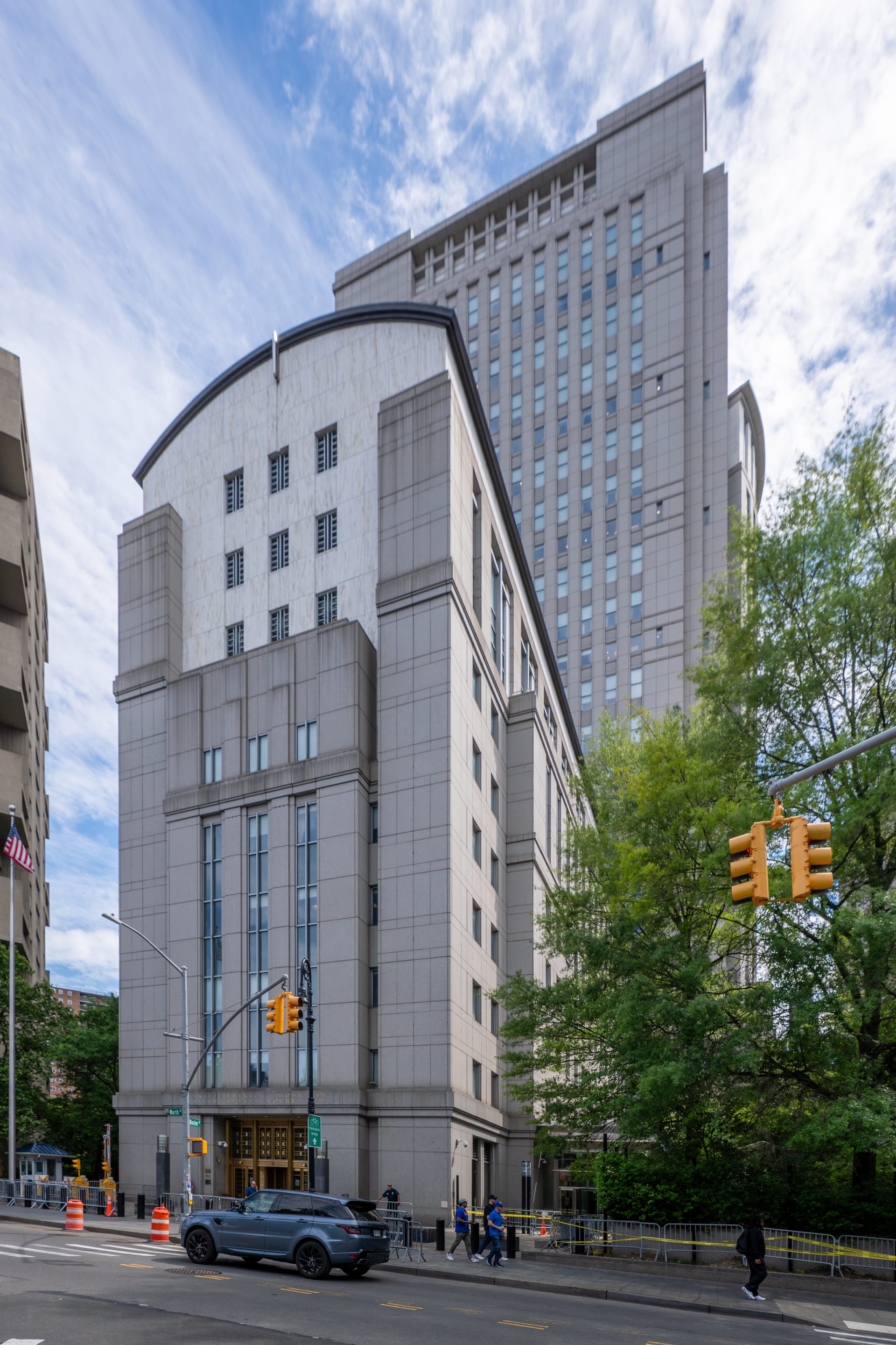 Gray granite facade of the federal courthouse in Manhattan, with a tall tower behind it, trees and a street with pedestrians.