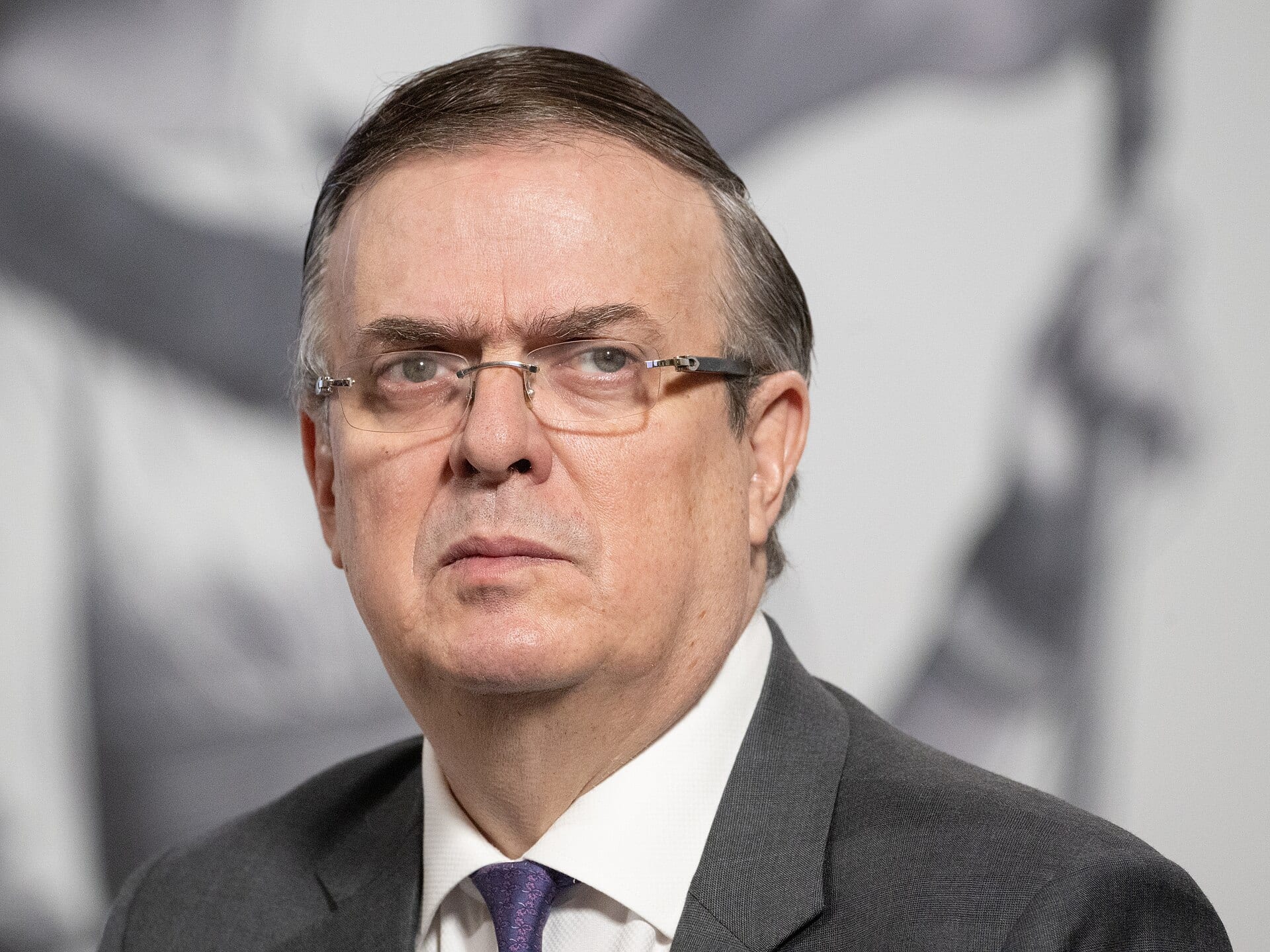 Retrato de un hombre con lentes sin marco, traje gris oscuro, camisa blanca y corbata morada, que mira hacia un lado con gesto serio.