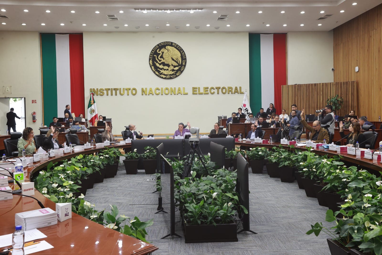 Consejeros y representantes de partidos levantan la mano para votar en el pleno del Consejo General, bajo el letrero del Instituto Nacional Electoral.