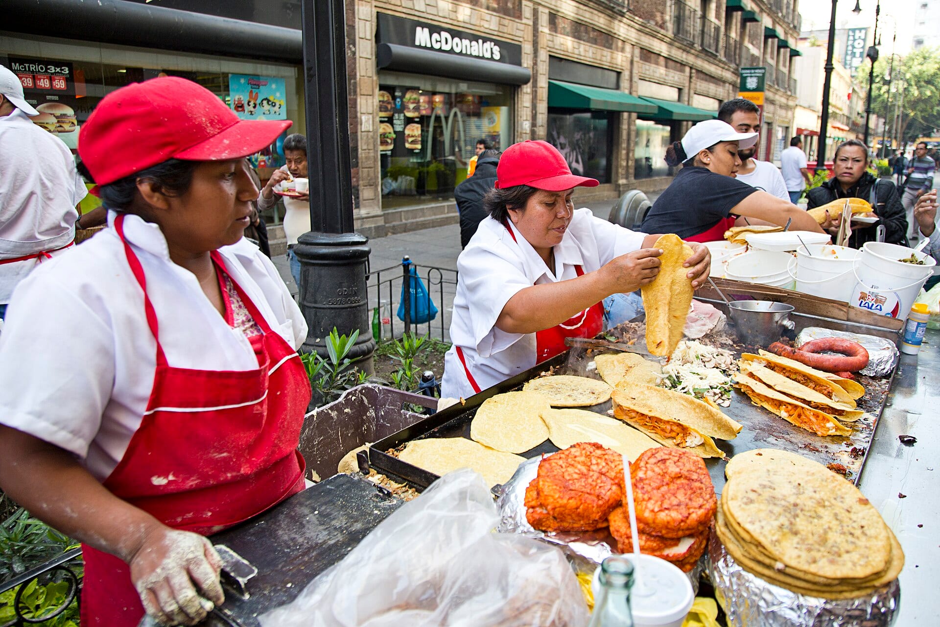 Dos mujeres con gorra roja y delantal preparan quesadillas y tacos en un puesto de comida callejera de una calle del centro de la Ciudad de México.