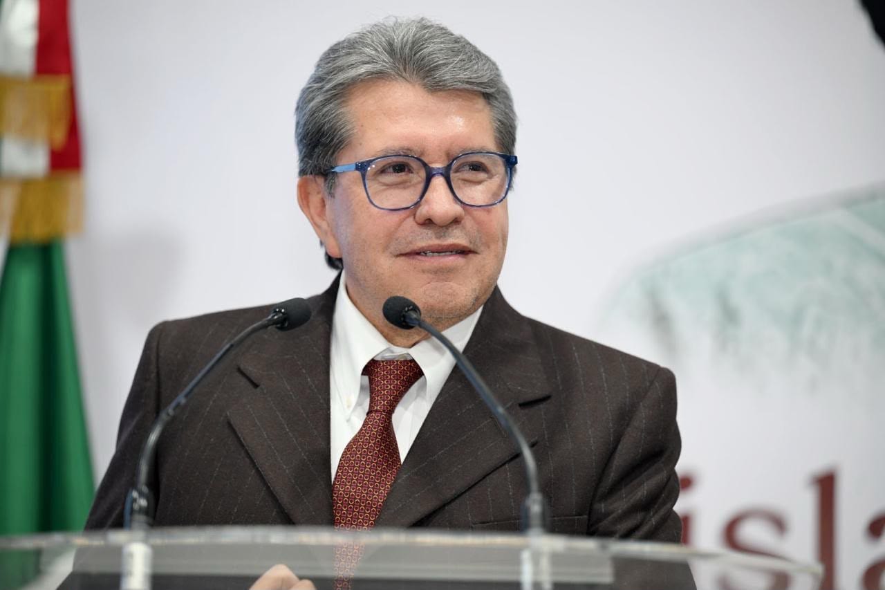 El diputado Ricardo Monreal sonríe detrás de dos micrófonos, en un podio de cristal, con una bandera de México a su izquierda.