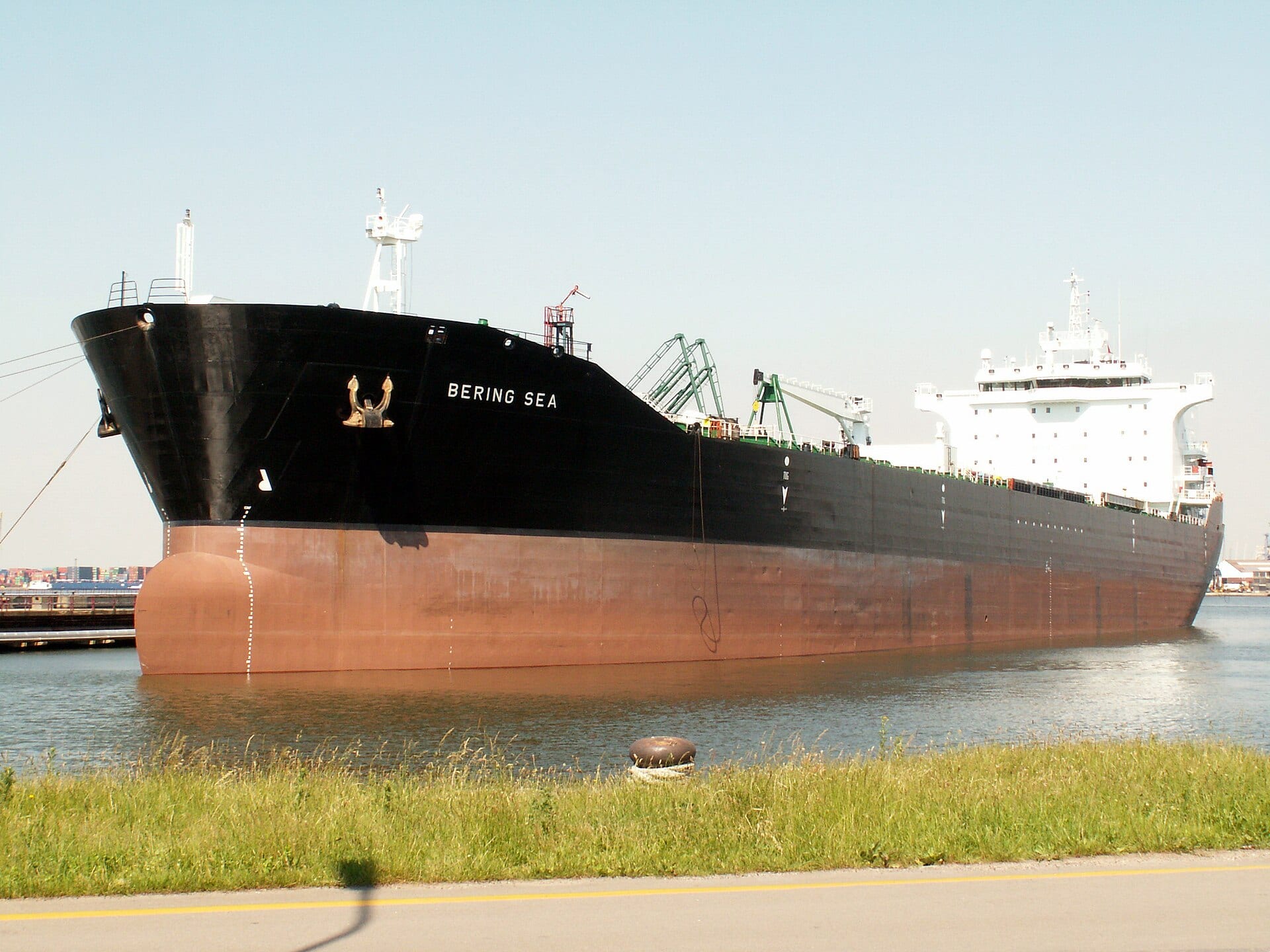 A black-and-red oil tanker moored at a port, with grass and a road in the foreground.