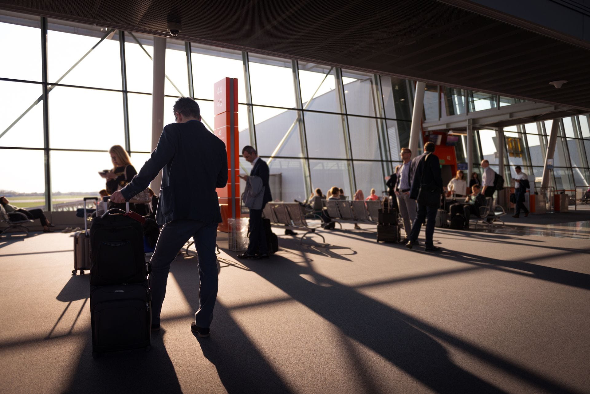 Un viajero con maleta de mano camina por una sala de espera de aeropuerto con grandes ventanales, rodeado de otros pasajeros.
