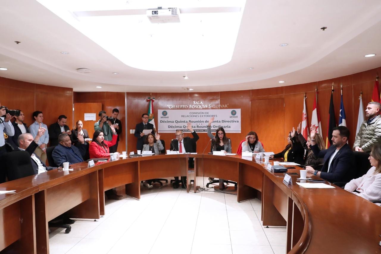 Diputados de la Comisión de Relaciones Exteriores levantan la mano para votar en una mesa semicircular, bajo una manta de la reunión, con fotógrafos al fondo.