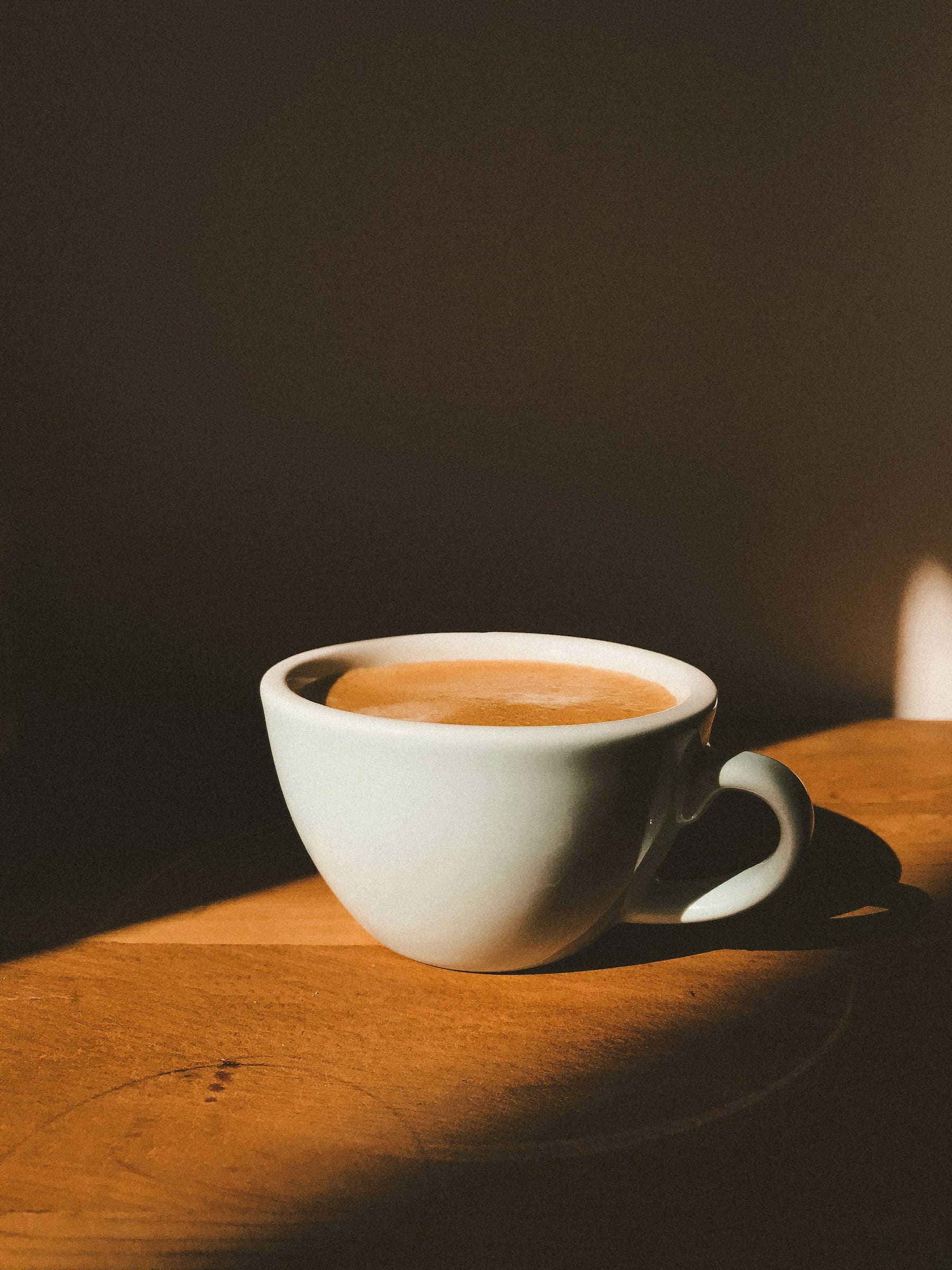 Taza blanca con café con espuma sobre una mesa de madera, con luz que entra de costado sobre un fondo oscuro.
