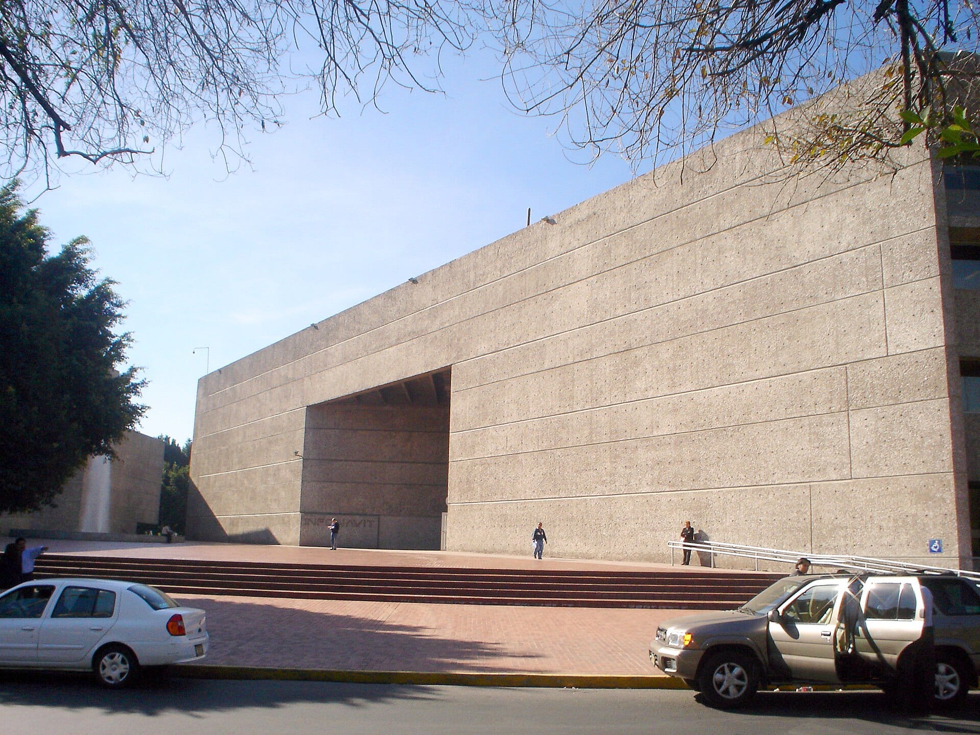 Fachada de concreto de la sede del Infonavit, con una explanada de ladrillo, escalones y varias personas frente al edificio.