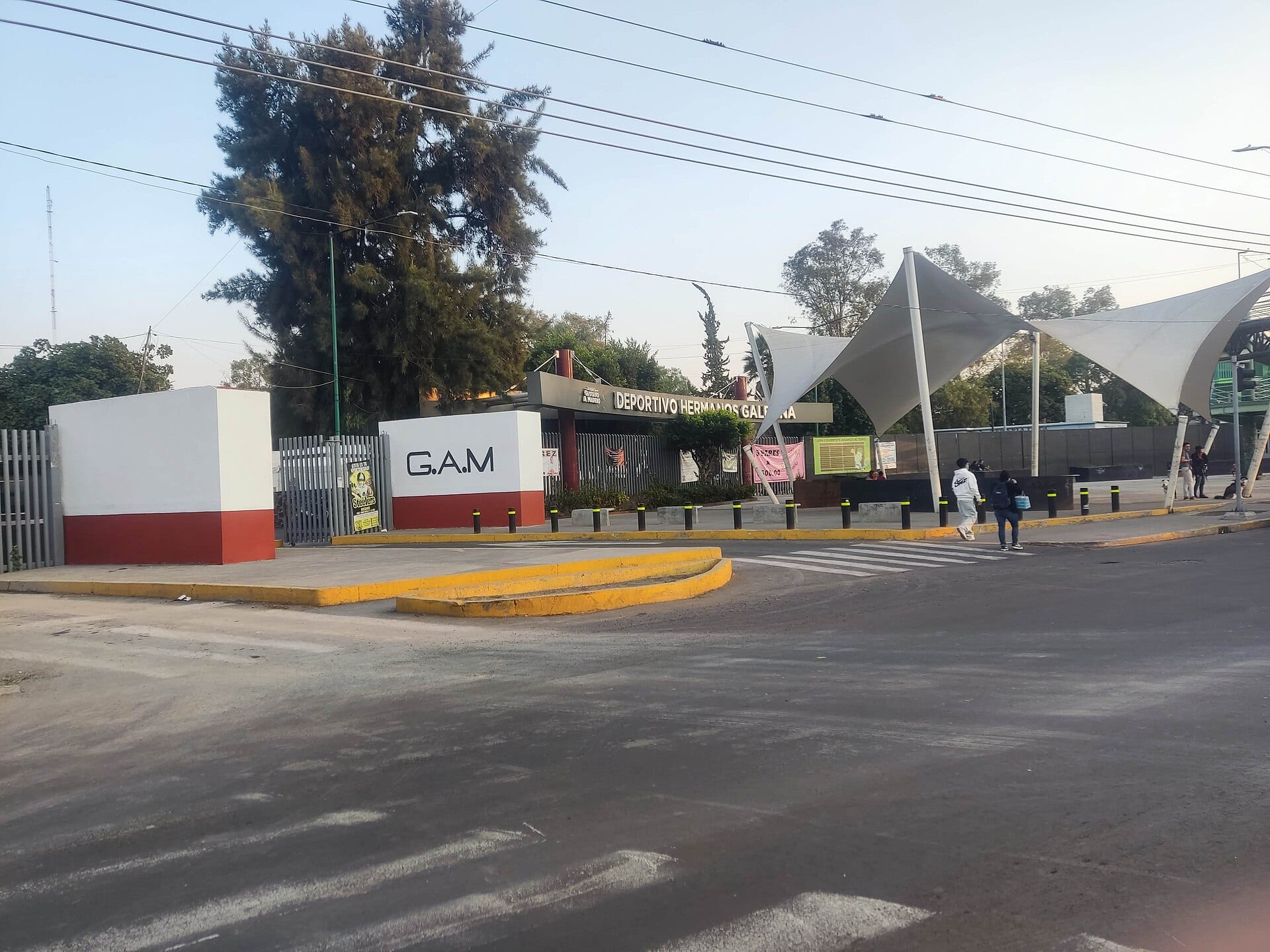 Entrada del Deportivo Hermanos Galeana, con un muro blanco y rojo con las letras G.A.M., una techumbre blanca de tela tensada y dos peatones cruzando la calle.