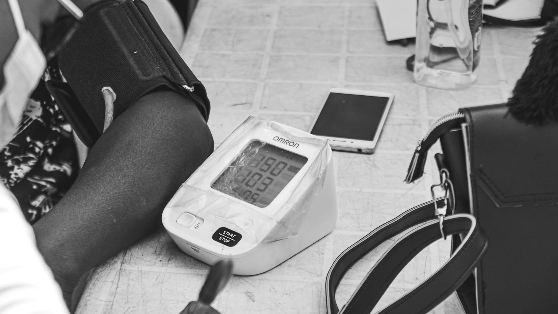 Brazo con brazalete negro junto a un monitor digital de presión arterial con pantalla encendida, un teléfono y un bolso sobre una mesa, en blanco y negro.