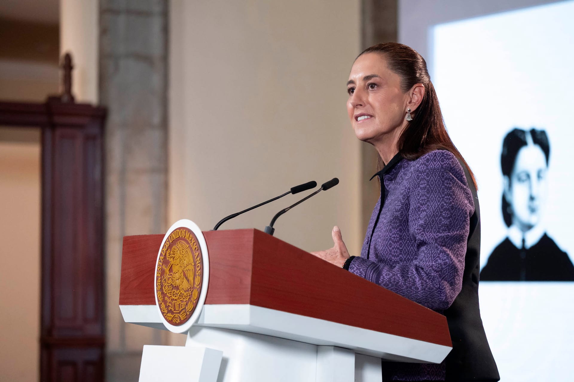 La presidenta Claudia Sheinbaum habla de perfil tras un podio con el escudo nacional, con una pantalla de fondo que muestra un retrato en blanco y negro.
