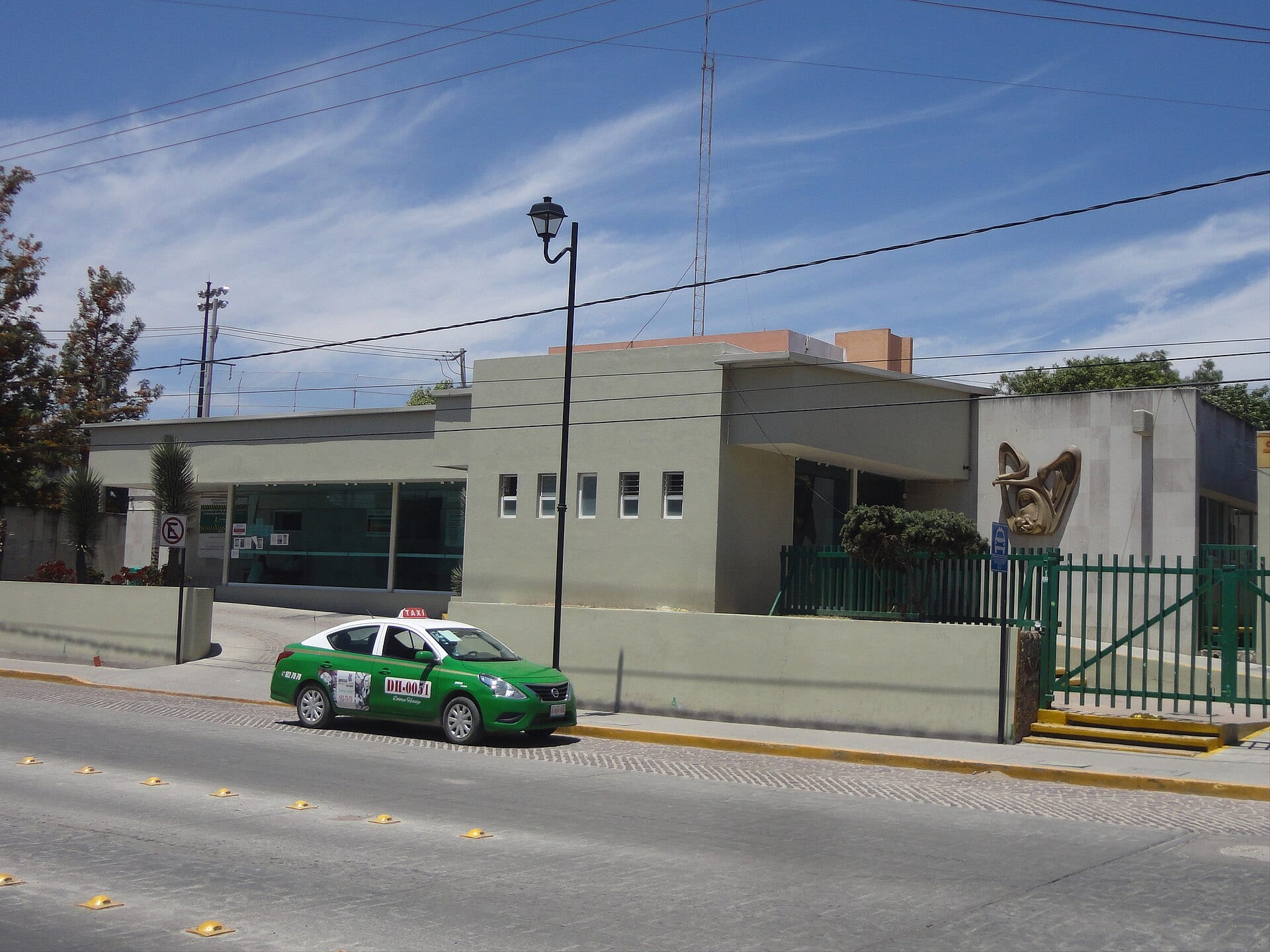 Fachada de una clínica del IMSS con el logotipo del instituto en el muro de la derecha y un taxi verde estacionado en la calle.