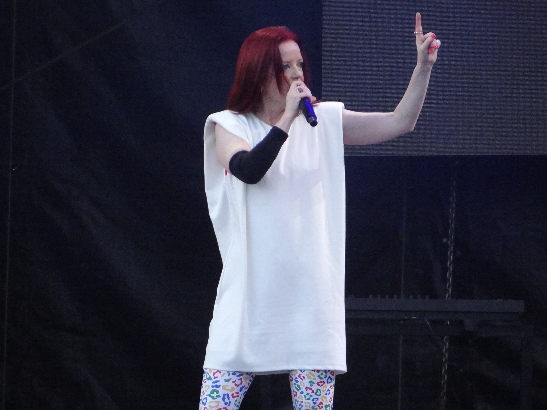 Una cantante de pelo rojo, con vestido blanco de hombreras y mallas de manchas de colores, canta con un micrófono y levanta una mano en un escenario oscuro.