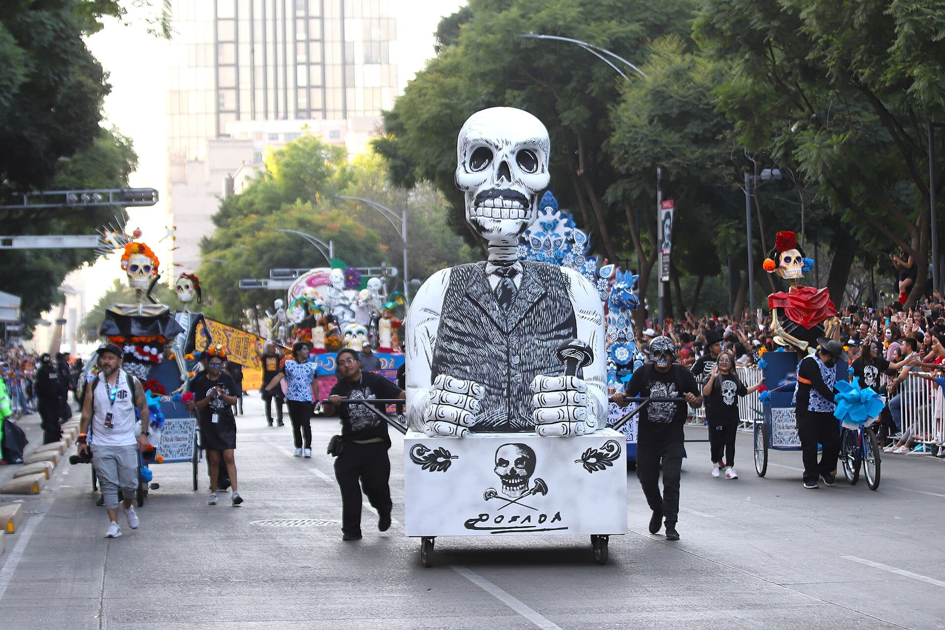 Una figura gigante de esqueleto con traje y bastón, firmada Posada, avanza sobre una plataforma que empujan dos personas por una avenida arbolada, entre público.