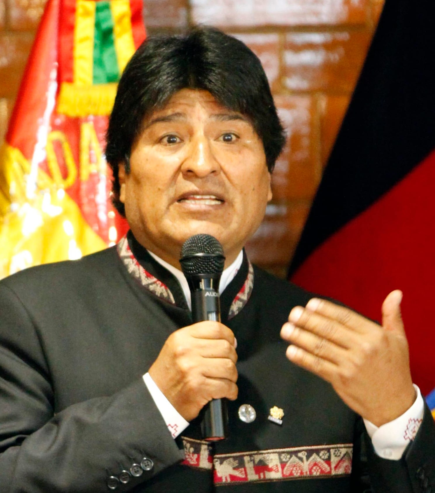 Un hombre habla ante un micrófono con la mano derecha en alto, vestido con chaqueta oscura de bordados andinos y banderas de fondo.