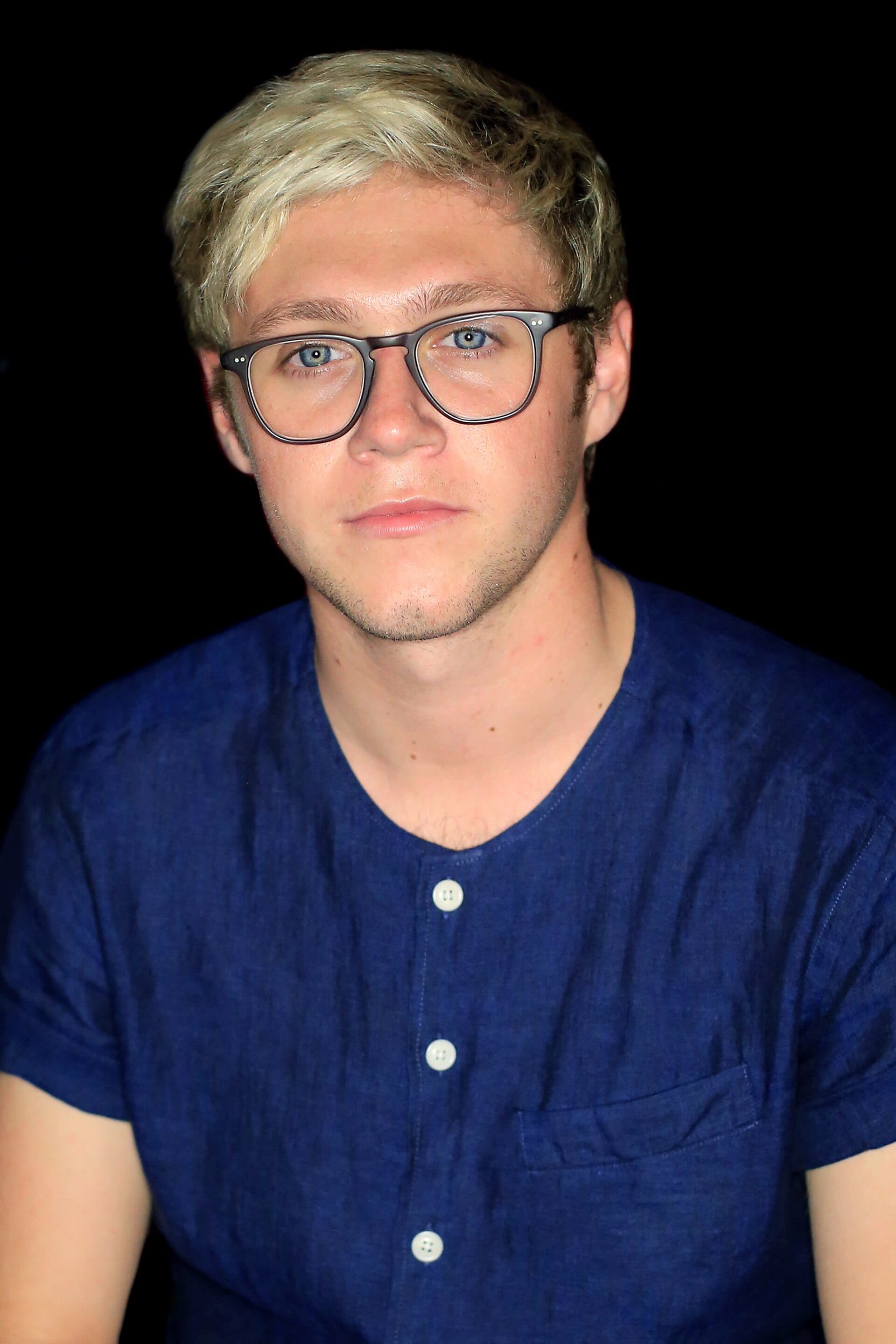 Niall Horan, de cabello rubio corto y lentes de armazón oscuro, con camisa azul marino, mira de frente sobre un fondo negro.