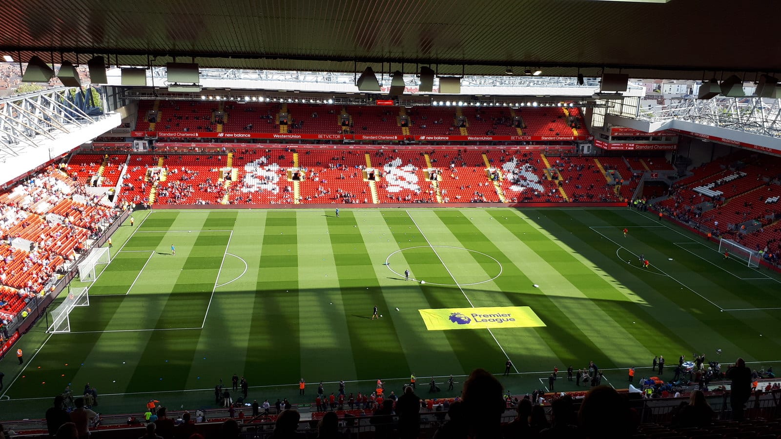 Vista general del estadio Anfield desde una grada alta, con el césped a rayas, tribunas rojas con pocos aficionados y una lona amarilla de la Premier League en el campo.