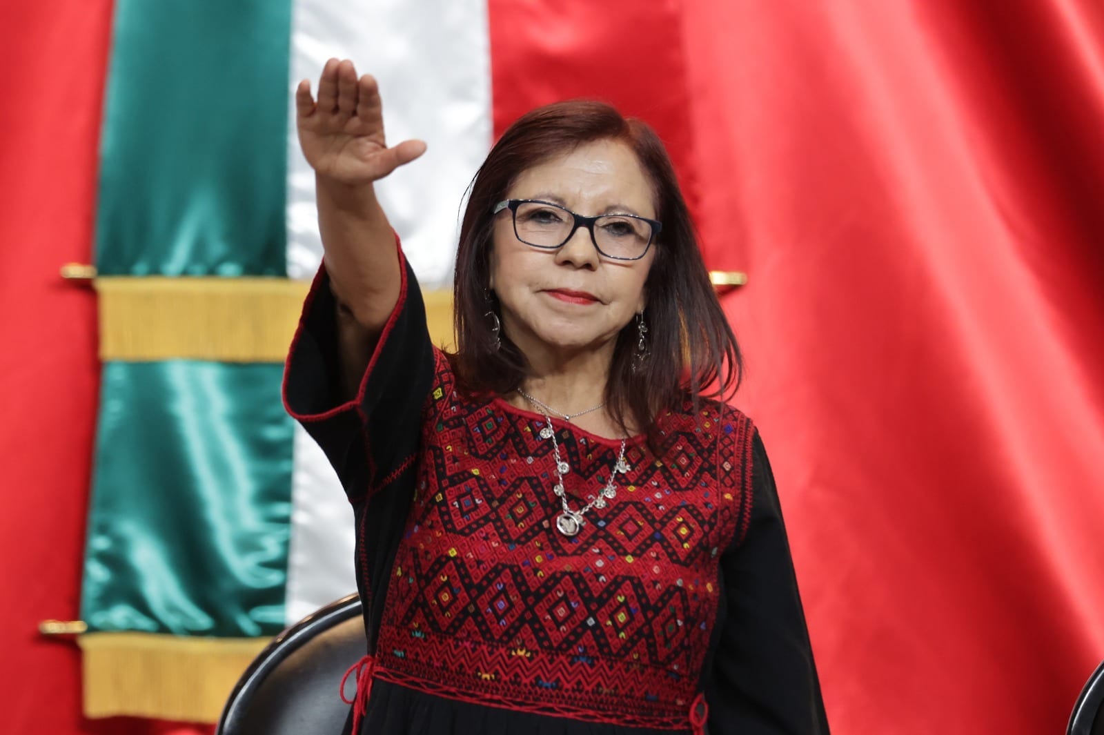 Leticia Ramírez Amaya, con lentes y huipil bordado en rojo y negro, levanta la mano derecha frente a una bandera de México.