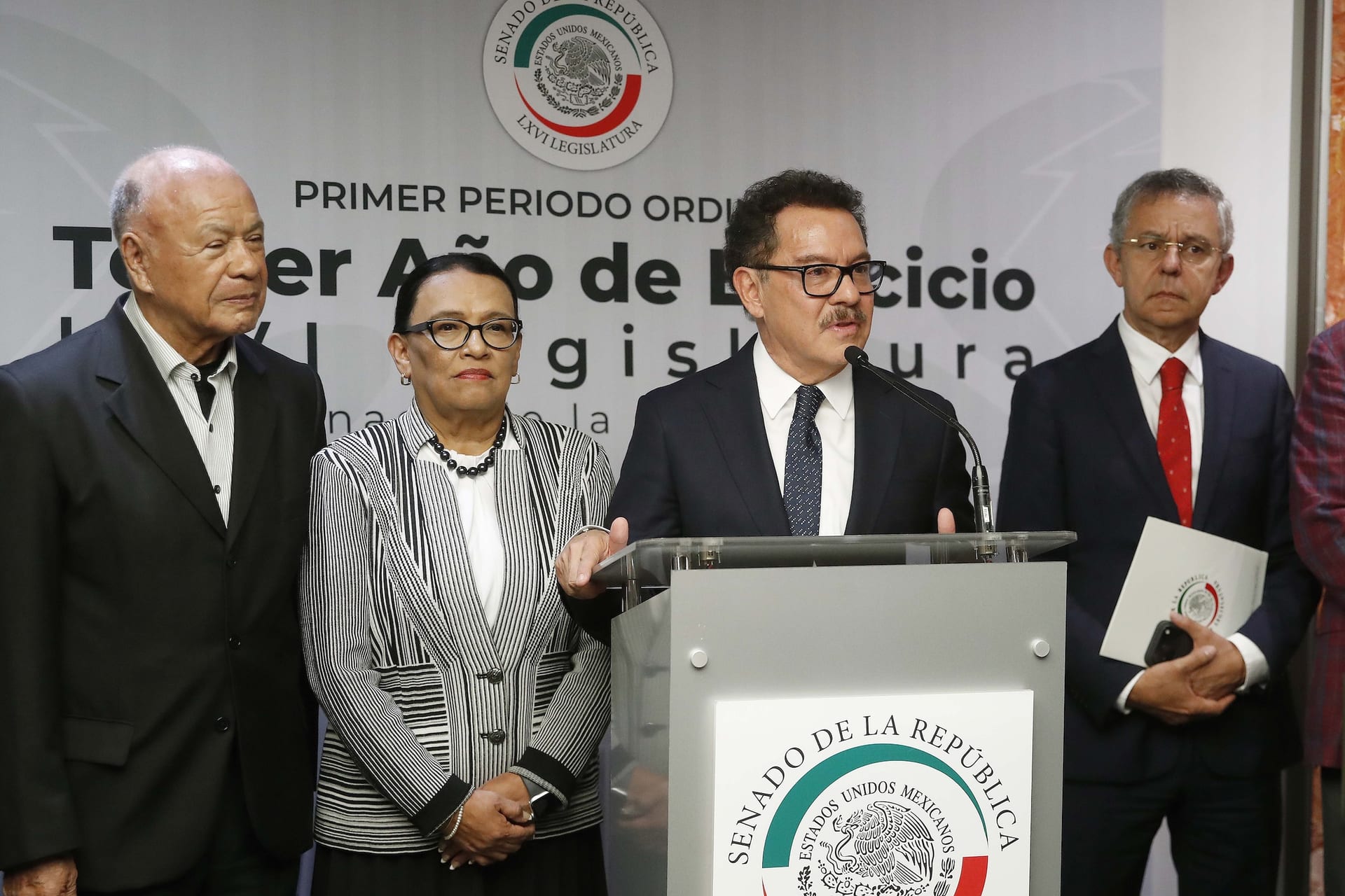 Rosa Icela Rodríguez, de saco a rayas blancas y negras, escucha a Ignacio Mier, que habla en un podio del Senado, junto a otros dos hombres.