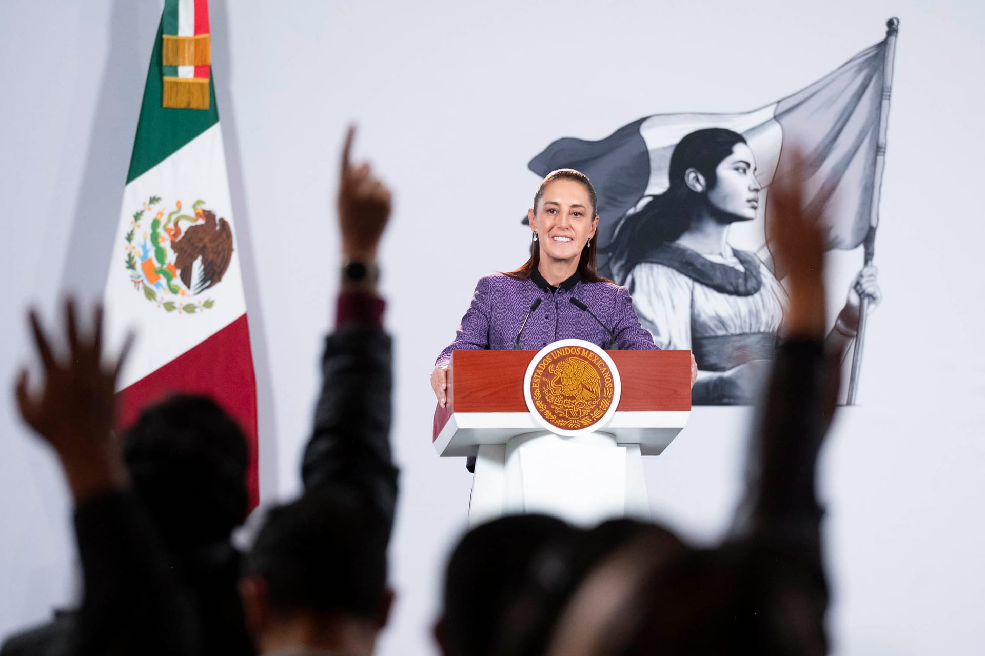 La presidenta Claudia Sheinbaum habla desde un podio con el escudo nacional mientras varios asistentes levantan la mano para pedir la palabra.