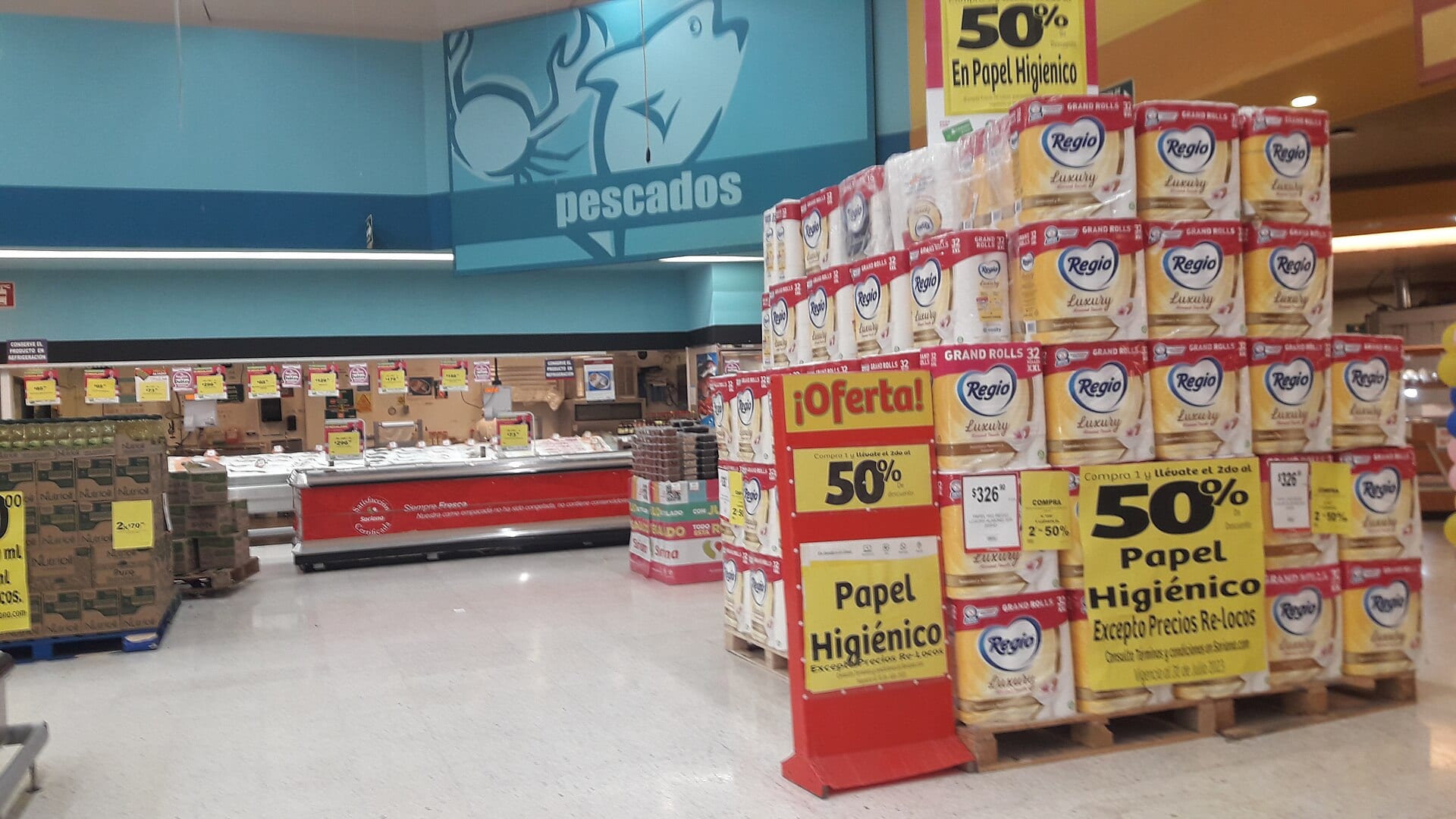Pasillo de un supermercado con una torre de paquetes de papel higiénico en oferta a la derecha y el mostrador de pescados al fondo.