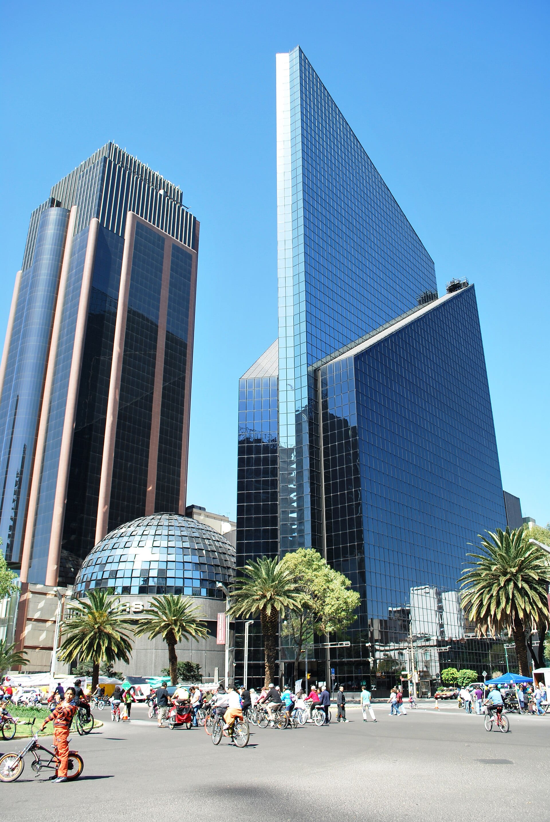 La cúpula de vidrio de la Bolsa Mexicana de Valores junto a dos torres de cristal, con ciclistas y peatones en la glorieta de Paseo de la Reforma.