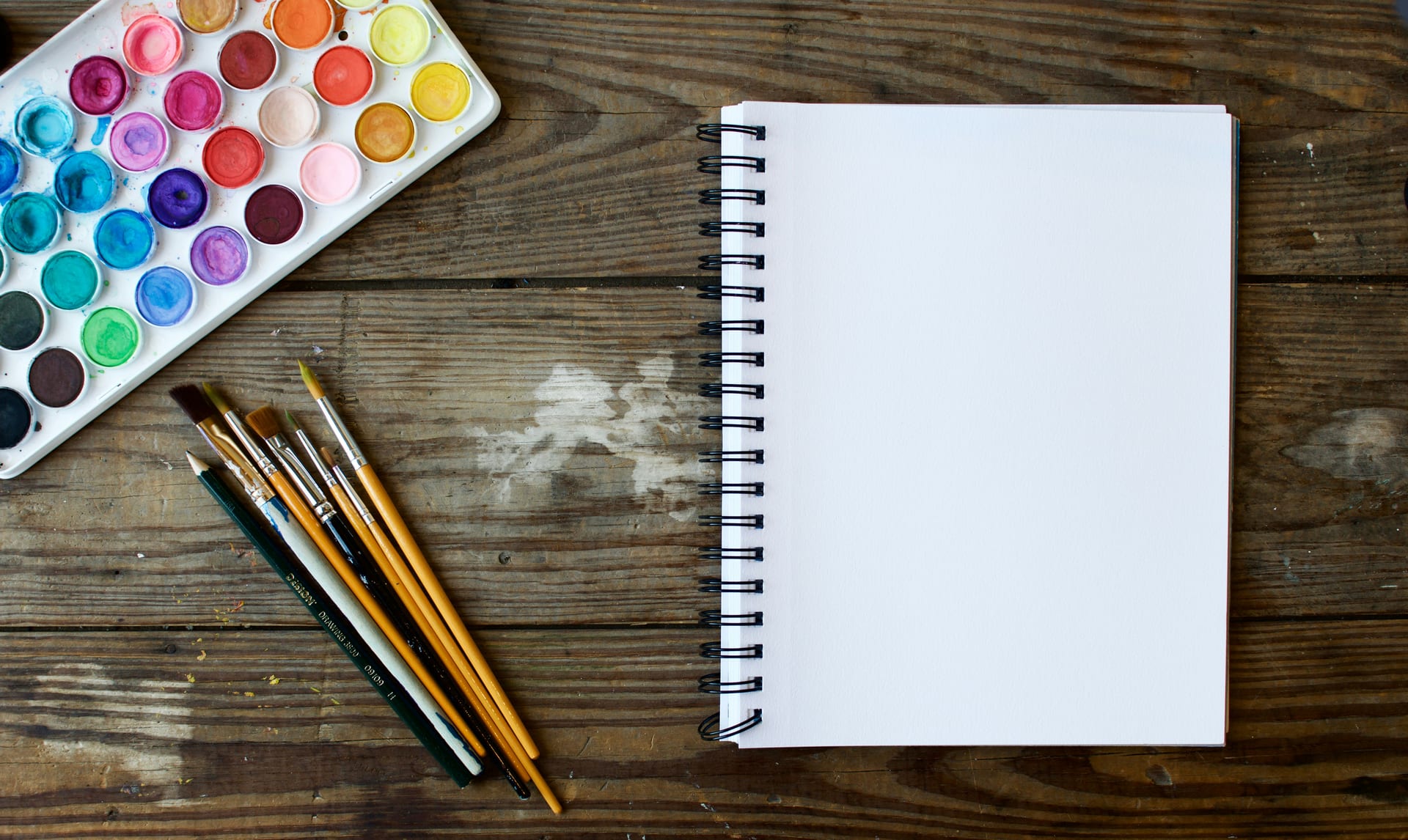 Pinceles junto a un cuaderno de dibujo en blanco y una paleta de acuarela sobre una mesa de madera.
