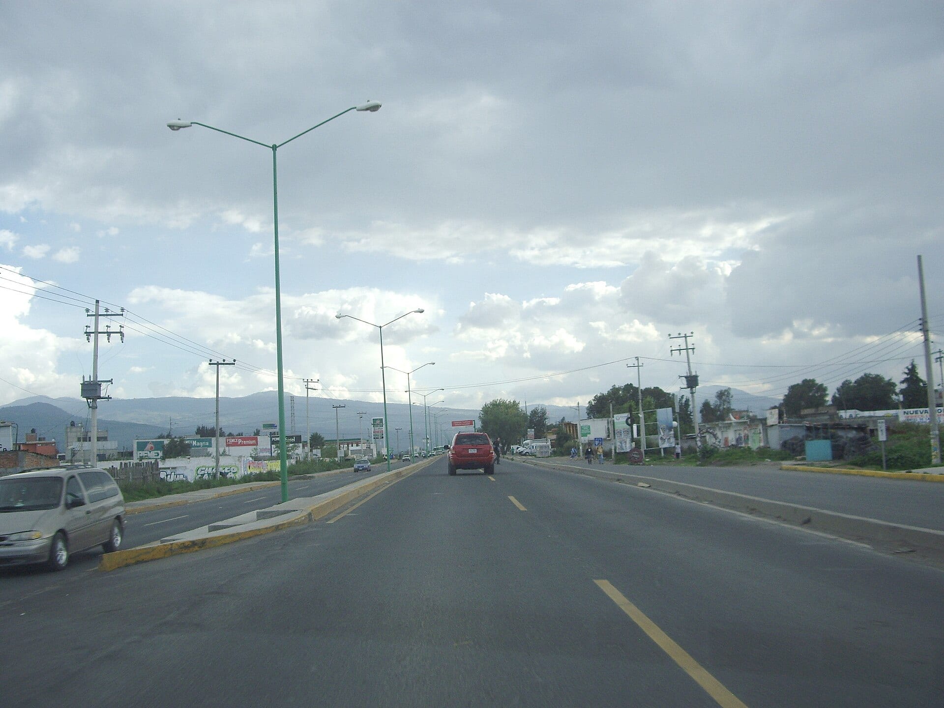 Una avenida de varios carriles con camellón amarillo, postes de alumbrado verdes y montañas al fondo bajo un cielo nublado.