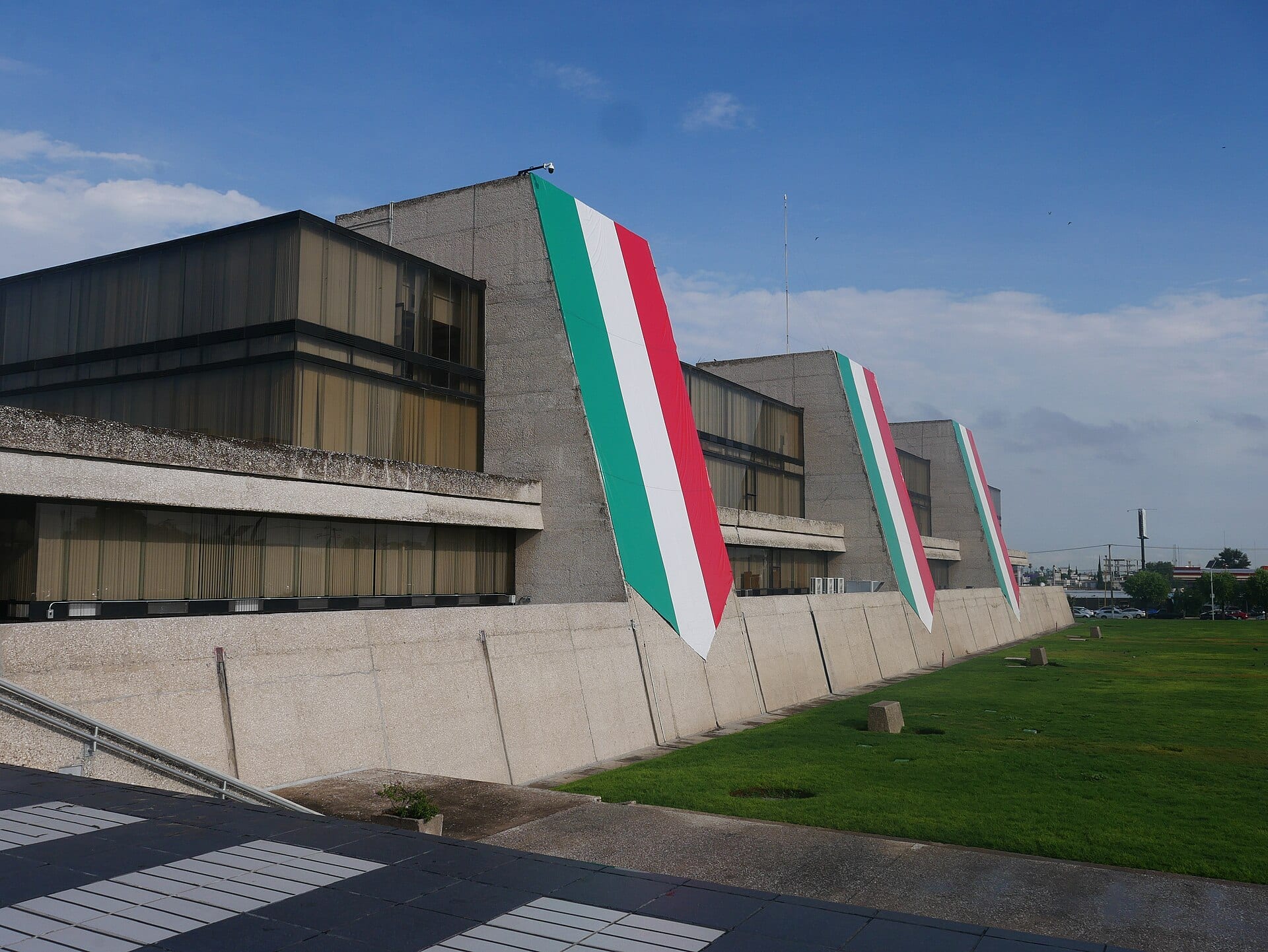 Costado de concreto del edificio central del INEGI, con tres banderas de México colgando de sus muros y césped al frente.