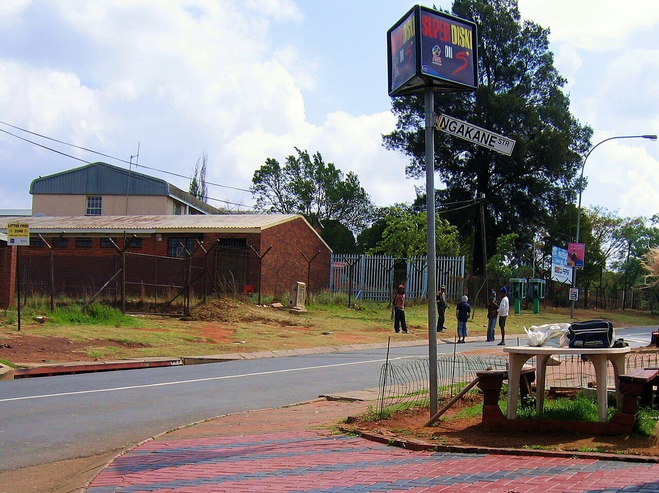 A street in Orlando West, Soweto, with a signpost and several people standing beside the sidewalk.