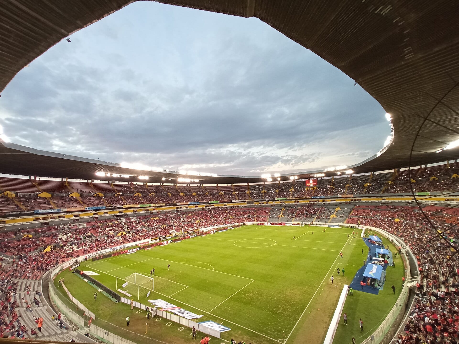Vista desde la tribuna alta del Estadio Jalisco, con la cancha de pasto verde, las gradas con aficionados de rojo y negro y el cielo nublado sobre el techo.
