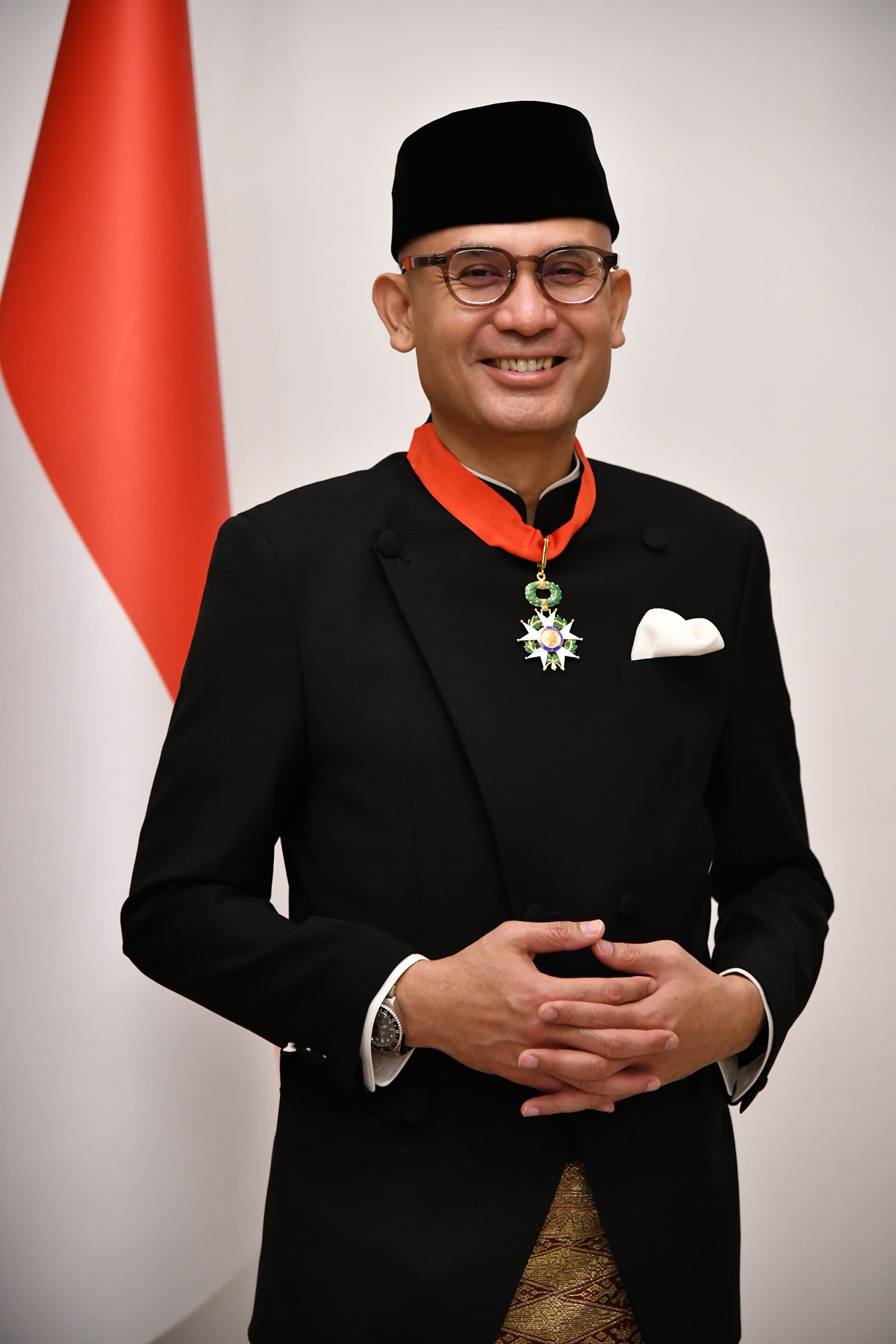 Arrmanatha Nasir sonríe con las manos entrelazadas, con traje negro, gorro negro y una condecoración al cuello, frente a la bandera roja y blanca de Indonesia.