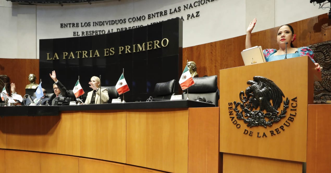 Una senadora levanta la mano desde el podio del pleno, con el escudo del Senado al frente, mientras otros legisladores votan a mano alzada en la mesa directiva.