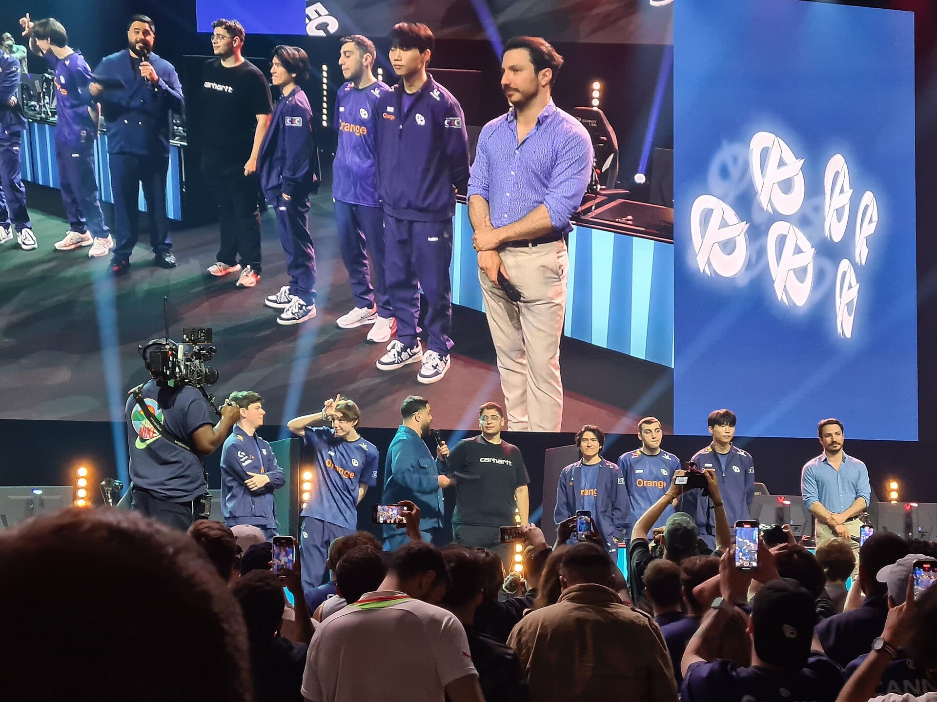 Karmine Corp players in blue and purple jackets standing on a stage in front of a giant screen with the team logo, as a crowd films on phones.
