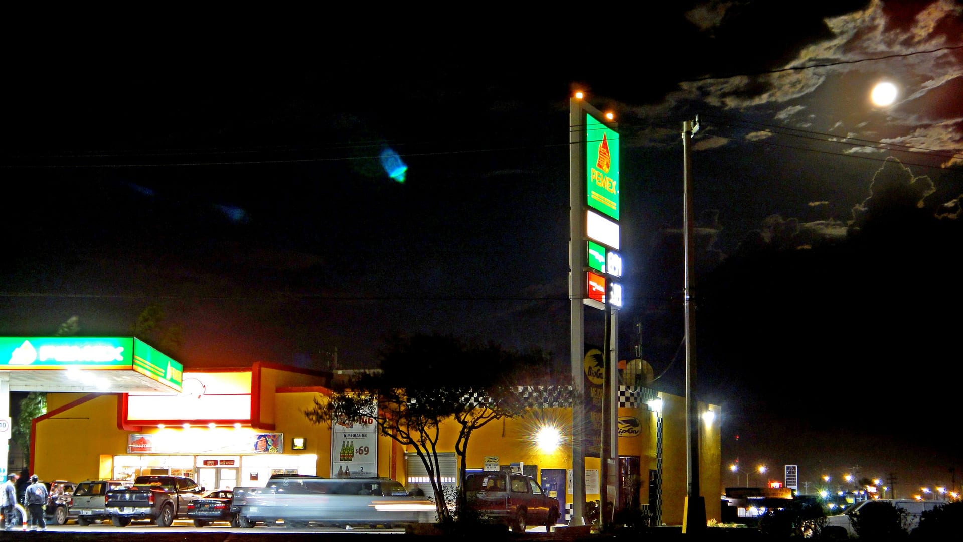 Una gasolinera Pemex iluminada durante la noche, con su letrero de precios y varios vehículos estacionados enfrente.