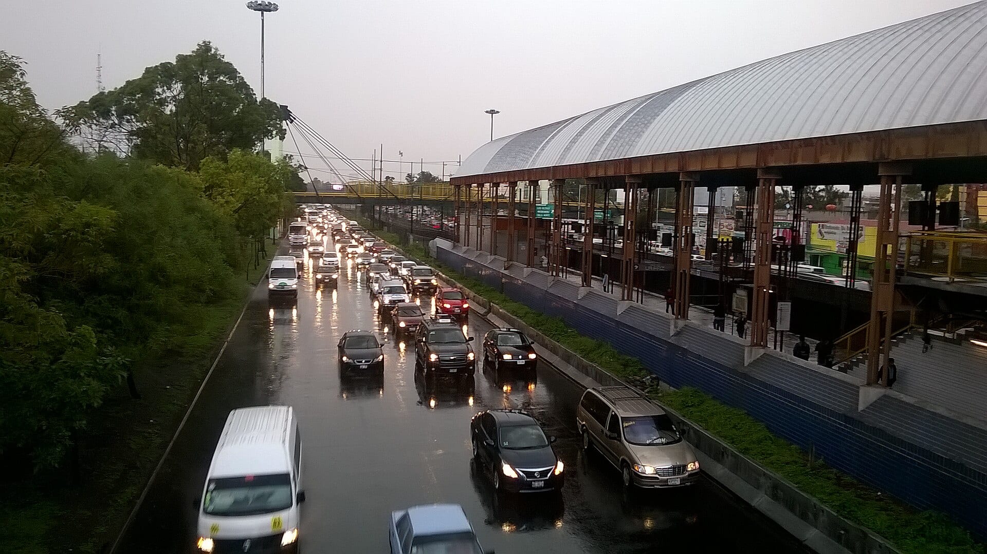 Autos con las luces encendidas avanzan despacio por una calzada mojada junto a una estación techada, bajo un cielo gris.