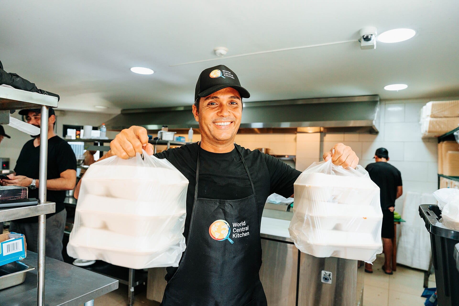 Un cocinero con delantal de World Central Kitchen sostiene dos bolsas con porciones de comida en una cocina.