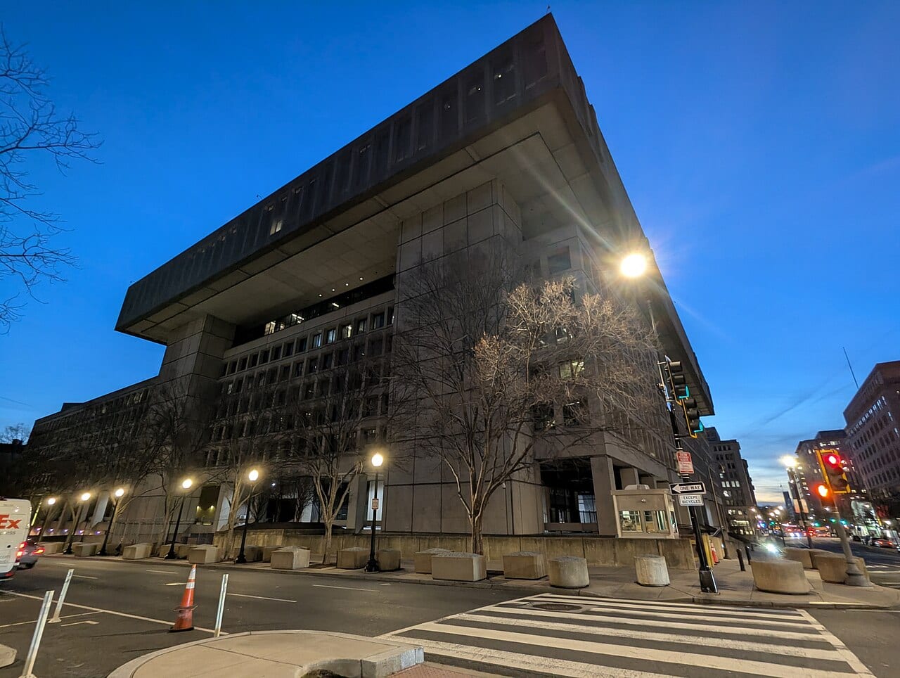 Concrete FBI headquarters building at dusk, with street lamps lit and a pedestrian crossing in the foreground.