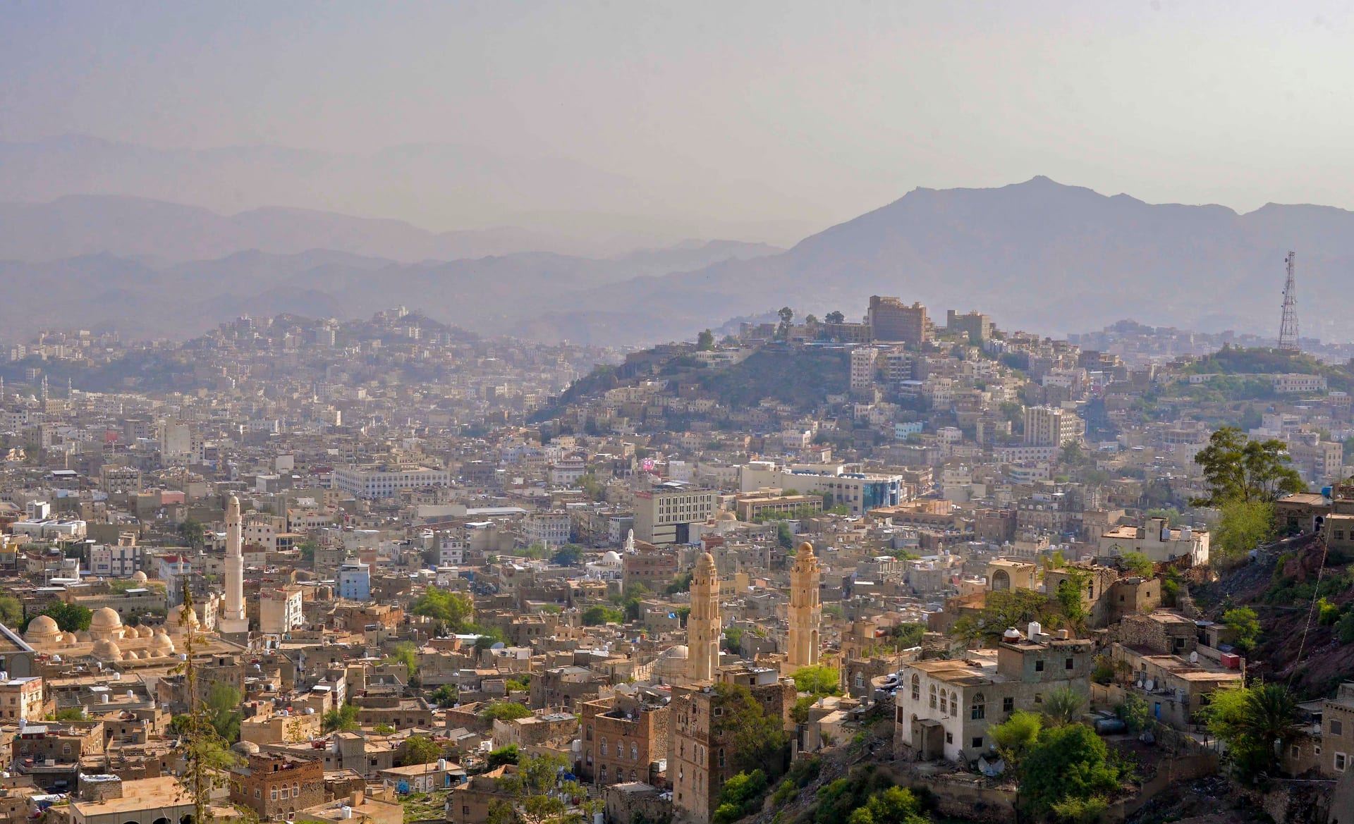 Panoramic view of the city of Taiz, with multi-story buildings climbing a mountainside under a hazy sky.