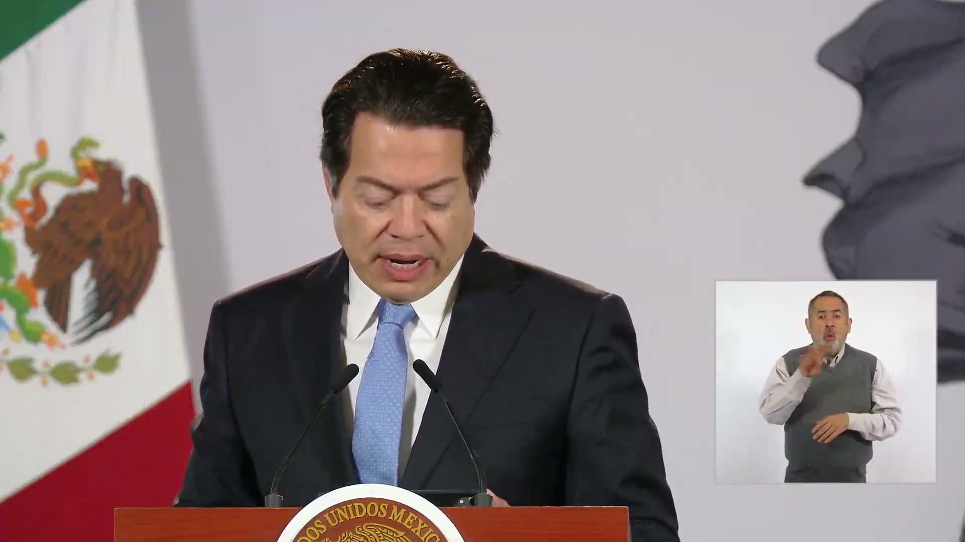 Un hombre de traje oscuro y corbata azul habla frente a un atril, con la bandera de México a su izquierda y un intérprete de lengua de señas a la derecha.