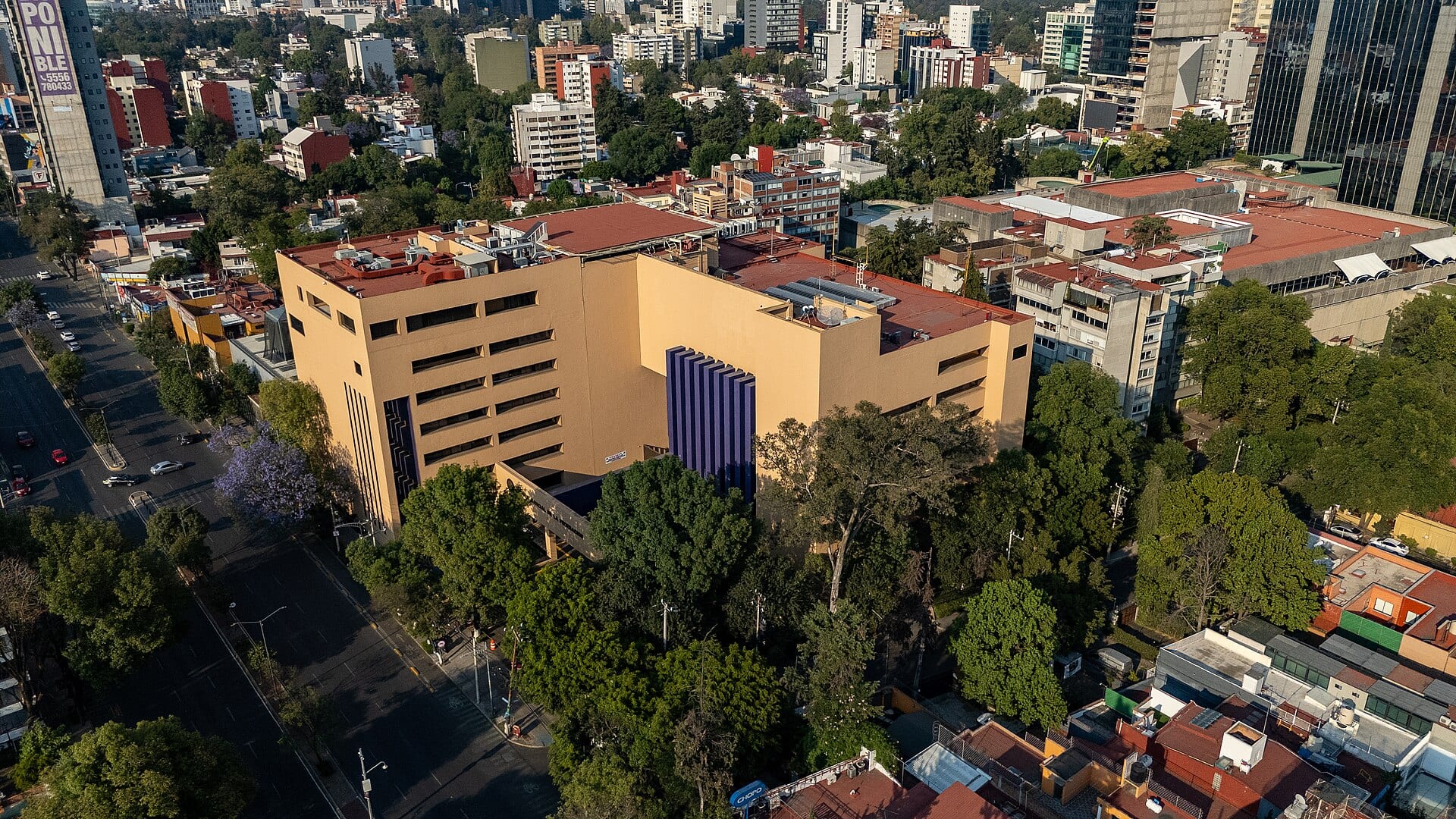 Vista aérea de un edificio de oficinas de color arena con una franja vertical morada, rodeado de árboles y calles de una colonia de la Ciudad de México.