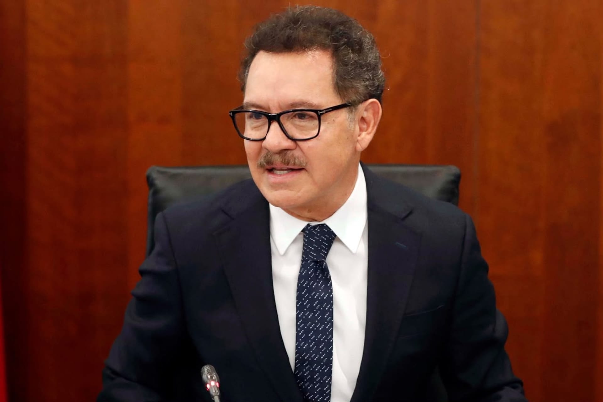 Un hombre de lentes de armazón negro y bigote, con traje oscuro y corbata azul, habla sentado frente a un micrófono ante un muro de madera.