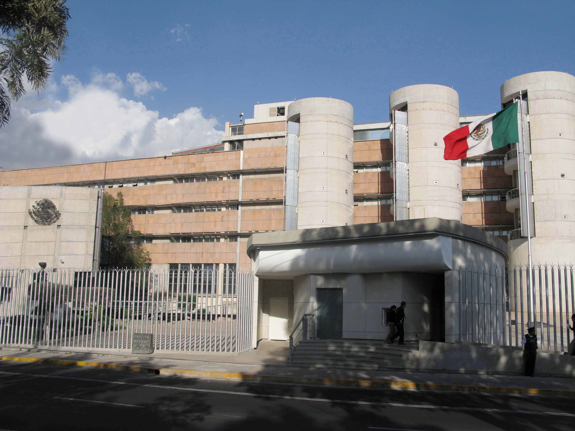 Edificio de concreto y tabique con tres torres cilíndricas, rodeado por una reja metálica, con una bandera de México ondeando a la derecha.